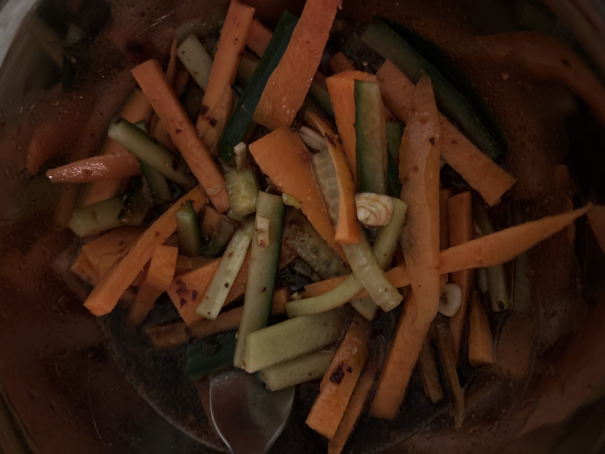 A top-down view shows a metal bowl containing sliced carrots and cucumbers in a liquid, possibly a marinade or dressing. The carrots are cut into rectangular sticks. The cucumbers, some still with the peel, are also cut into sticks. There's a utensil, possibly a spoon or fork, present in the lower left portion of the bowl. The contents of the bowl are surrounded by the dark background of the interior of the bowl, suggesting a dimly lit indoor setting. There is no indication this image was taken in London.