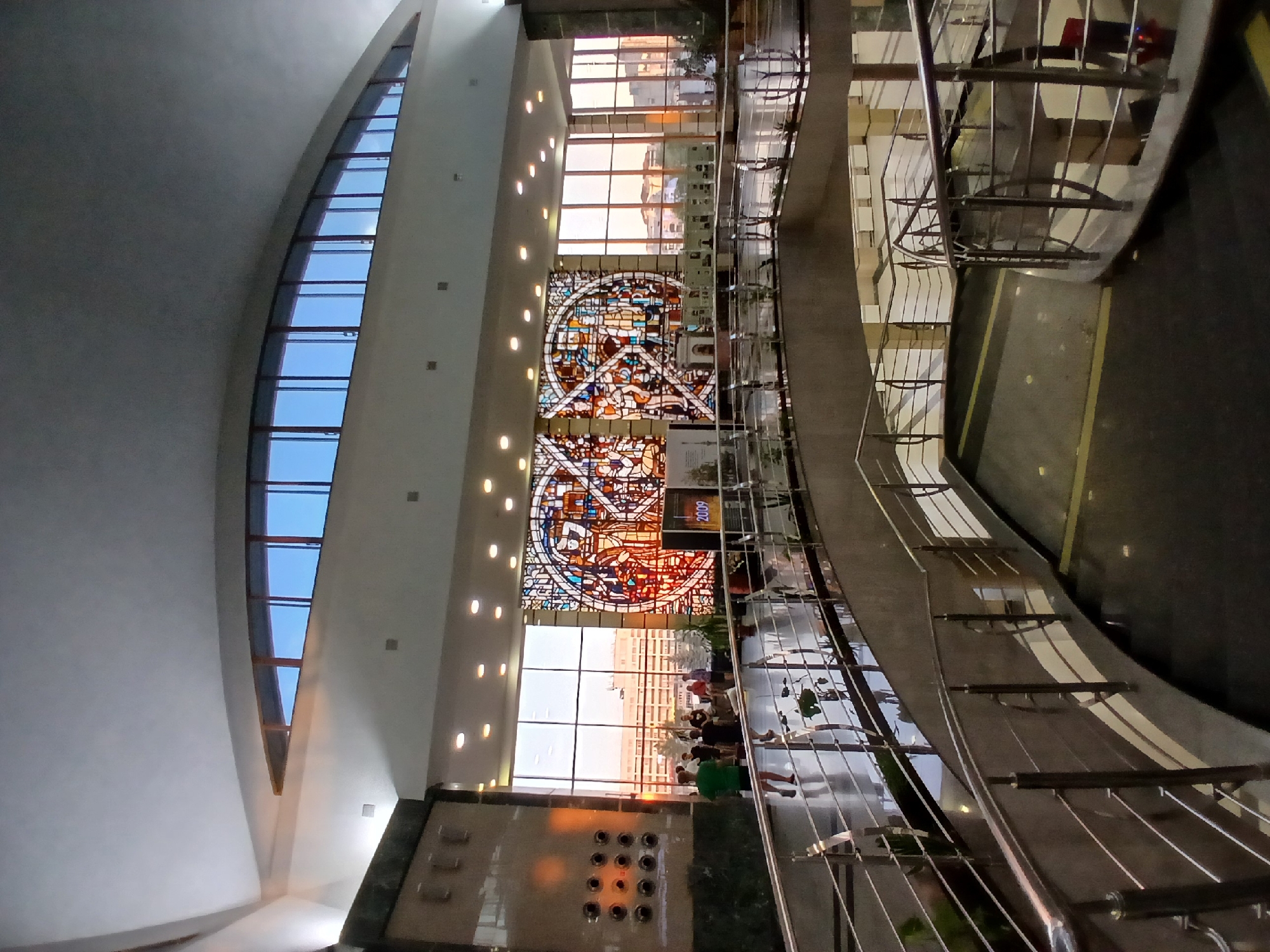 The image shows the interior of a large, modern building, likely a public space or exhibition hall, with a prominent stained-glass window and a curved ramp or walkway in the foreground. The stained-glass artwork is colorful and features geometric patterns and possibly figures. Large windows behind the stained glass reveal an urban landscape with buildings bathed in the warm light of what appears to be sunset or sunrise. In the mid-ground, several people are standing and conversing near the stained-glass window. The foreground features a metallic railing and a dark, polished floor that reflects the surrounding light and structures. The architecture includes a high, arched ceiling with recessed lighting. The scene is likely the Chelyabinsk State Museum of Fine Arts, known for its stained-glass art.