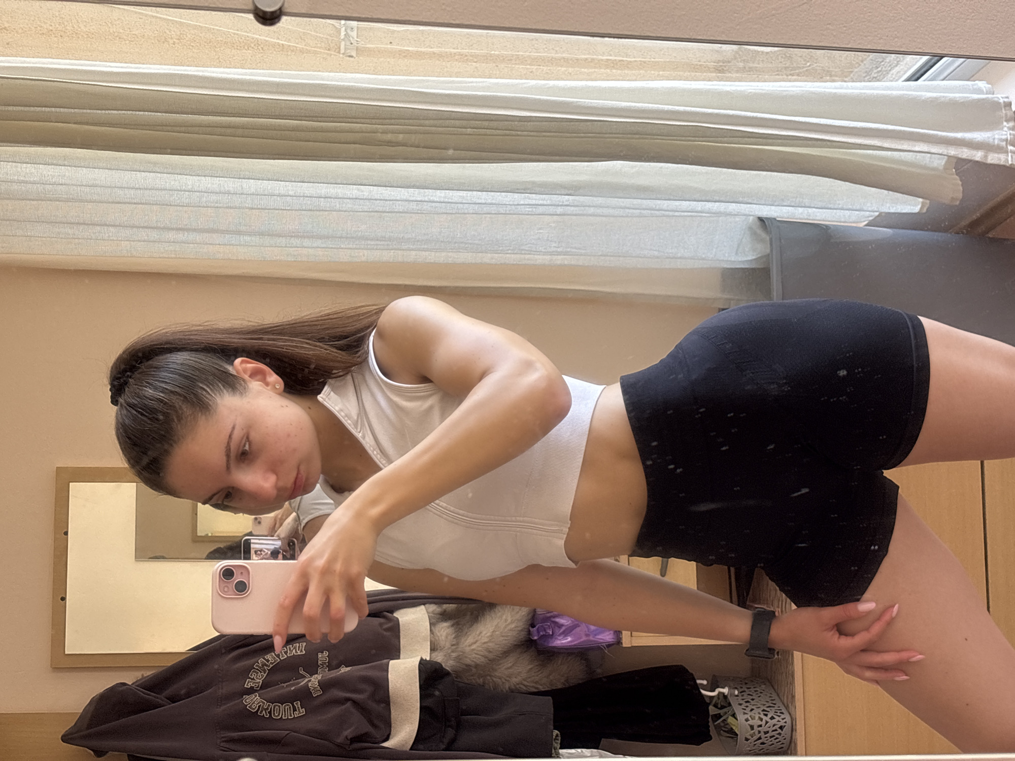 A young female, appearing to be in her early twenties with light skin, dark hair tied in a high ponytail with a black scrunchie, and a small gold stud earring in her left ear, is captured in a mirror selfie within an indoor setting in Varna, Bulgaria. She is holding a light pink smartphone with visible dual cameras in her left hand, positioned to take the photo, and has light pink nail polish on her visible fingers. She wears a dark-colored smartwatch on her left wrist. Her expression is neutral, and her gaze is directed towards the phone screen.
She is dressed in activewear, consisting of a white, sleeveless, cropped top with a partial front zipper and a hood, revealing her midriff. The top has visible ribbed textures and seams. She is also wearing high-waisted, black, short athletic shorts, also with a textured finish. Her right arm is slightly bent, and her left hand rests on her left thigh.
The background reflects an interior room, likely a bedroom or dressing area, with light beige walls. To the left of the subject, a dark brown/black jacket with a lighter trim and mirrored text (partially legible as "SENSE" and "RUNT") hangs from a hook or rail. Below the jacket, a garment or accessory with brown and grey fur is visible, alongside a small purple plastic bag or container. Further to the left and lower, a light grey woven basket filled with small items sits on a light wooden dresser or cabinet. Above the jacket, a smaller, rectangular mirror with a light wooden frame hangs on the wall, showing further reflections of the room.
To the right of the subject, a large window is visible, covered by vertical, light beige or off-white blinds or sheer curtains that are drawn, allowing diffuse light into the room. Through the upper right portion of the window, a textured, light-colored building exterior or wall is partially visible, indicating an urban or residential outdoor environment. A dark grey bin or container is partially reflected in the bottom right corner of the mirror.