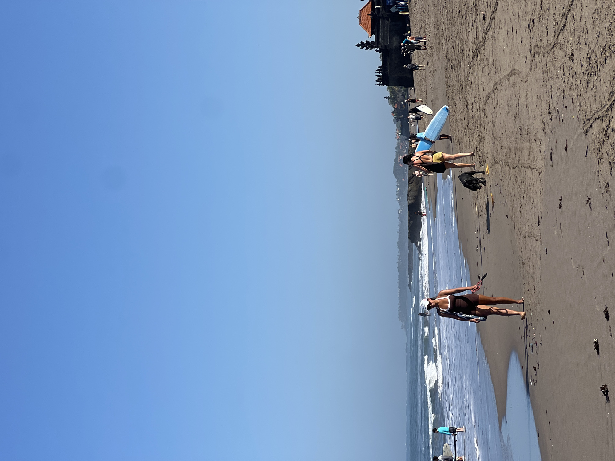 The image depicts a beach scene in North Kuta, Indonesia. The sand is dark, and the ocean has moderate waves. Several people are present on the beach and in the water. In the foreground, a woman in a brown swimsuit and white hat walks along the wet sand, holding a surfboard and a leash attached to a black dog. Behind her, two women walk with surfboards, one carrying a light blue surfboard. A black dog is near them, sniffing at the sand. In the middle ground, other individuals can be seen walking and standing on the beach, some with surfboards. The water shows whitecaps from the waves. In the background, a large, ornate dark stone structure with a reddish-orange roof, possibly a temple or historical building, is visible on a slight incline. The sky is clear and blue. The overall scene suggests a typical day at a popular beach destination.