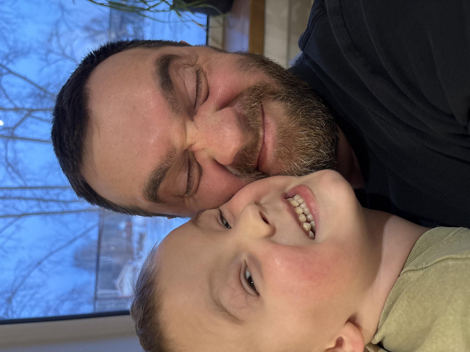 A man and a child are in a close, affectionate embrace. The man has a beard and dark hair and is wearing a dark shirt. His eyes are closed and he appears to be grinning. The child is lying on their back, also grinning, and is wearing a light green shirt. They are indoors, with a window showing a snowy landscape visible in the background. Although this photo was found in a location described as Chaikovsky, Russia, there is no way to verify this from visual data alone.