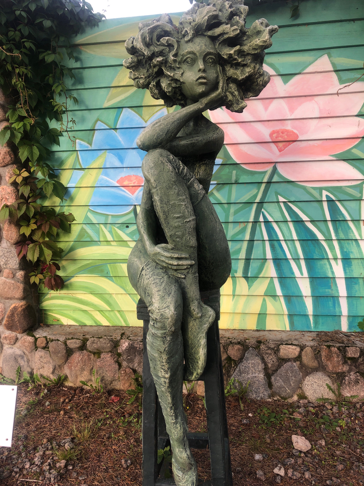 The image shows a bronze statue of a woman with a large, wild head of hair, seated on a dark stool. The statue is green and appears weathered. Behind the statue is a wooden wall painted with a mural of large pink and blue lotus flowers and green leaves. To the left of the statue, a stone wall is visible, covered in green ivy. The ground in front of the statue is covered in dirt, pine needles, and small stones. A white sign is partially visible on the ground to the left. The scene is outdoors, with natural light suggesting daytime. The location is identified as Интурист Снт, Russia.