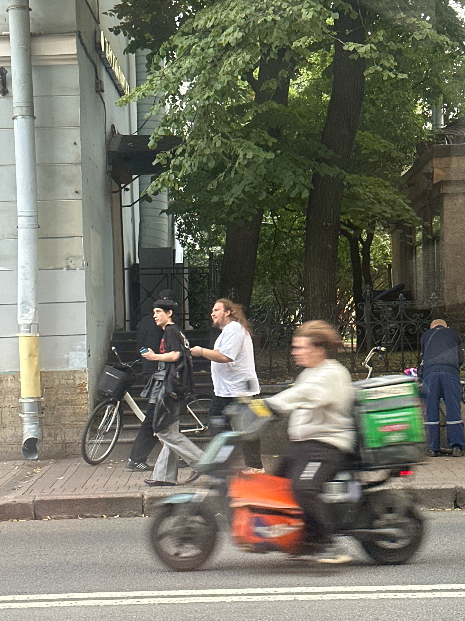 The image captures a street scene in Saint Petersburg, Russia. In the foreground, a person on a motorized scooter is blurred due to motion, wearing a light-colored jacket and dark pants. The scooter has a large green insulated bag mounted on the back and an orange storage compartment on the front. To the left of the scooter, on the sidewalk, two individuals are walking. One is wearing a black t-shirt, black pants, and a black hat, carrying a black bag over their shoulder. The other person, to their right, is wearing a white t-shirt and dark pants. A bicycle is parked behind these two individuals. The background shows a building with a pale facade and architectural details, surrounded by trees with lush green foliage. Further to the right, a person in a dark blue uniform with reflective stripes is standing near a wrought-iron fence. The sidewalk is paved with bricks, and a street runs alongside it.