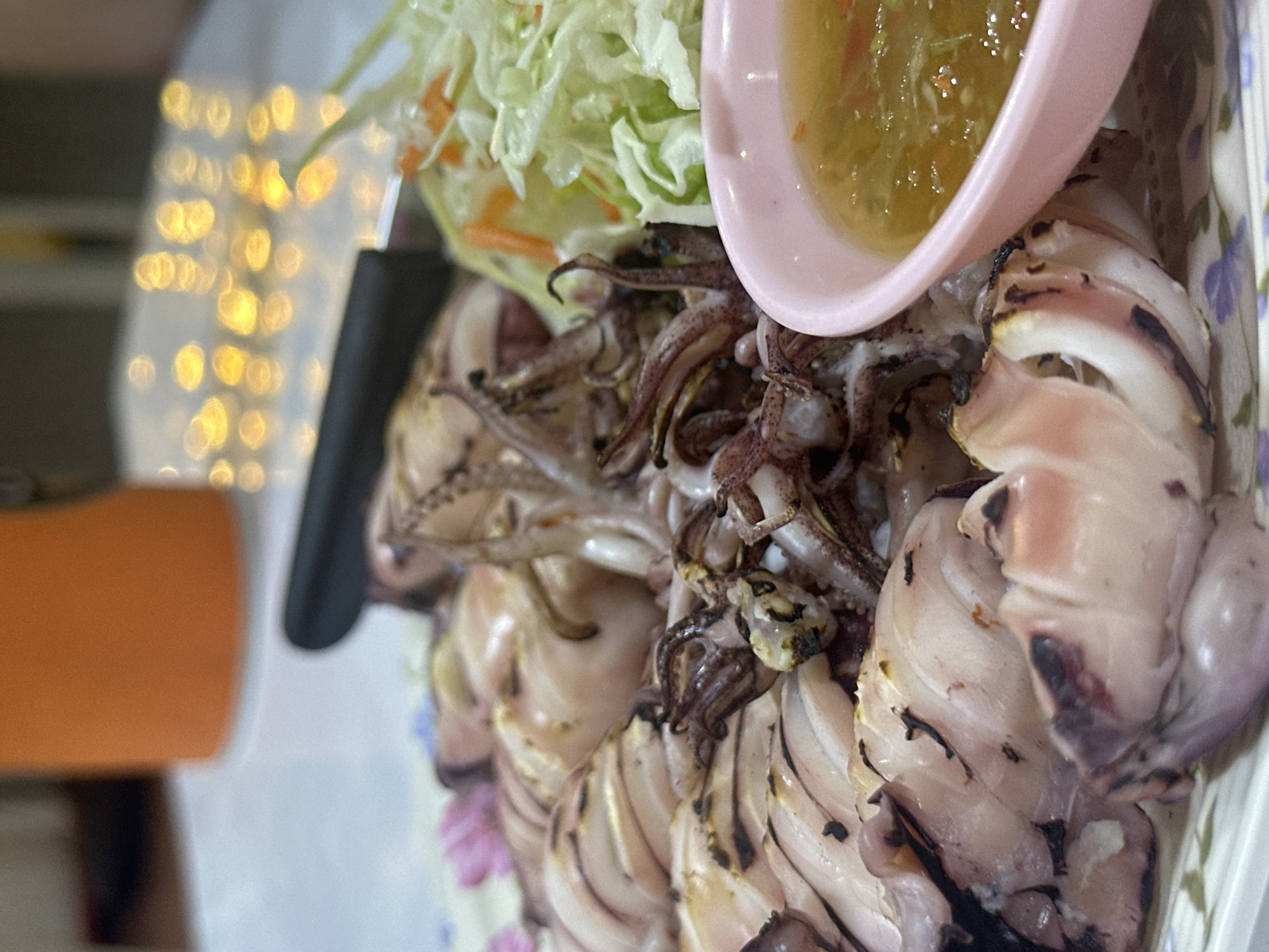 This close-up shot displays a plate of grilled squid served with a small pink dish of dipping sauce and a side of shredded cabbage and carrots. The squid, sliced and slightly charred, sits prominently on a plate decorated with a floral pattern. A black-handled knife rests on the plate near the squid and cabbage. The background is out of focus, showing blurred lights, possibly from a nearby establishment.
The location is assumed to be Pattaya City, Thailand, based on image context clues.