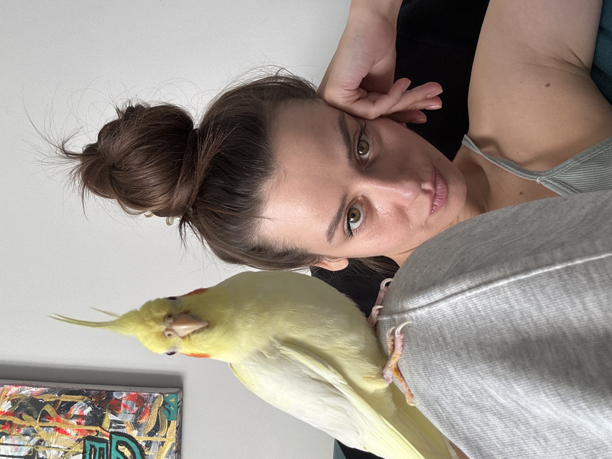 A woman with brown hair styled in a bun is shown from a close-up, canted angle. She rests her head on her hand and gazes directly at the viewer. A yellow cockatiel perches on her shoulder. A piece of abstract art with visible brushstrokes is visible in the lower left corner of the frame. There is no evidence in this image to suggest this photograph was taken in the city of Krasnoyarsk, Russia.