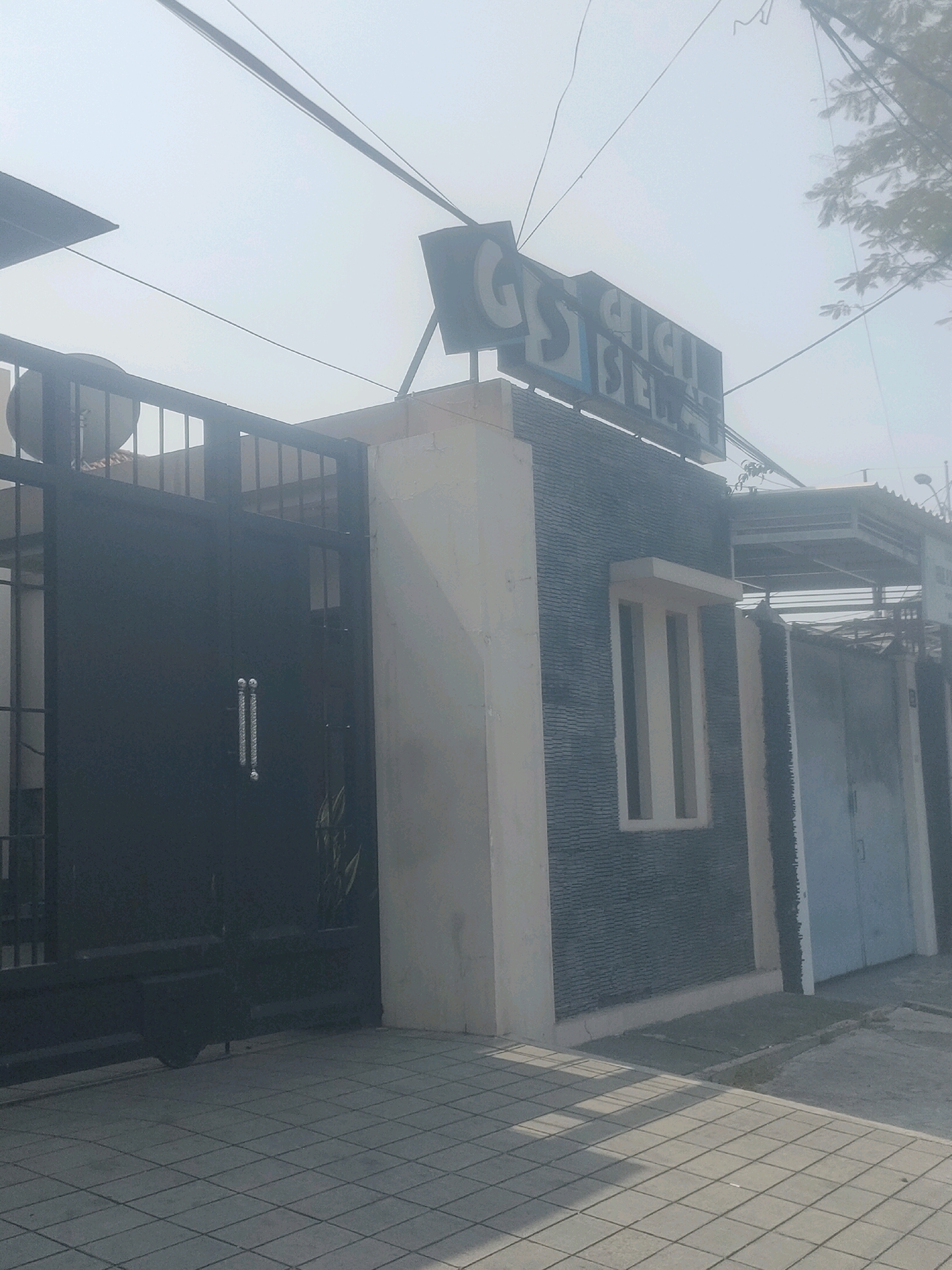 An outdoor, eye-level, slightly low-angle full shot shows the exterior of a building in Surabaya, Indonesia, on a hazy day. The building is modern, with a dark gray tiled facade and a beige section. A large, black metal gate is visible on the left side of the image, with two vertical silver handles. Above the building, a sign with the letters "GS GAJA SEJA" is visible against the bright, overcast sky. Power lines crisscross the sky. To the right of the tiled building, a light blue metal door or gate is partially visible. The ground in front of the building is paved with square gray tiles.