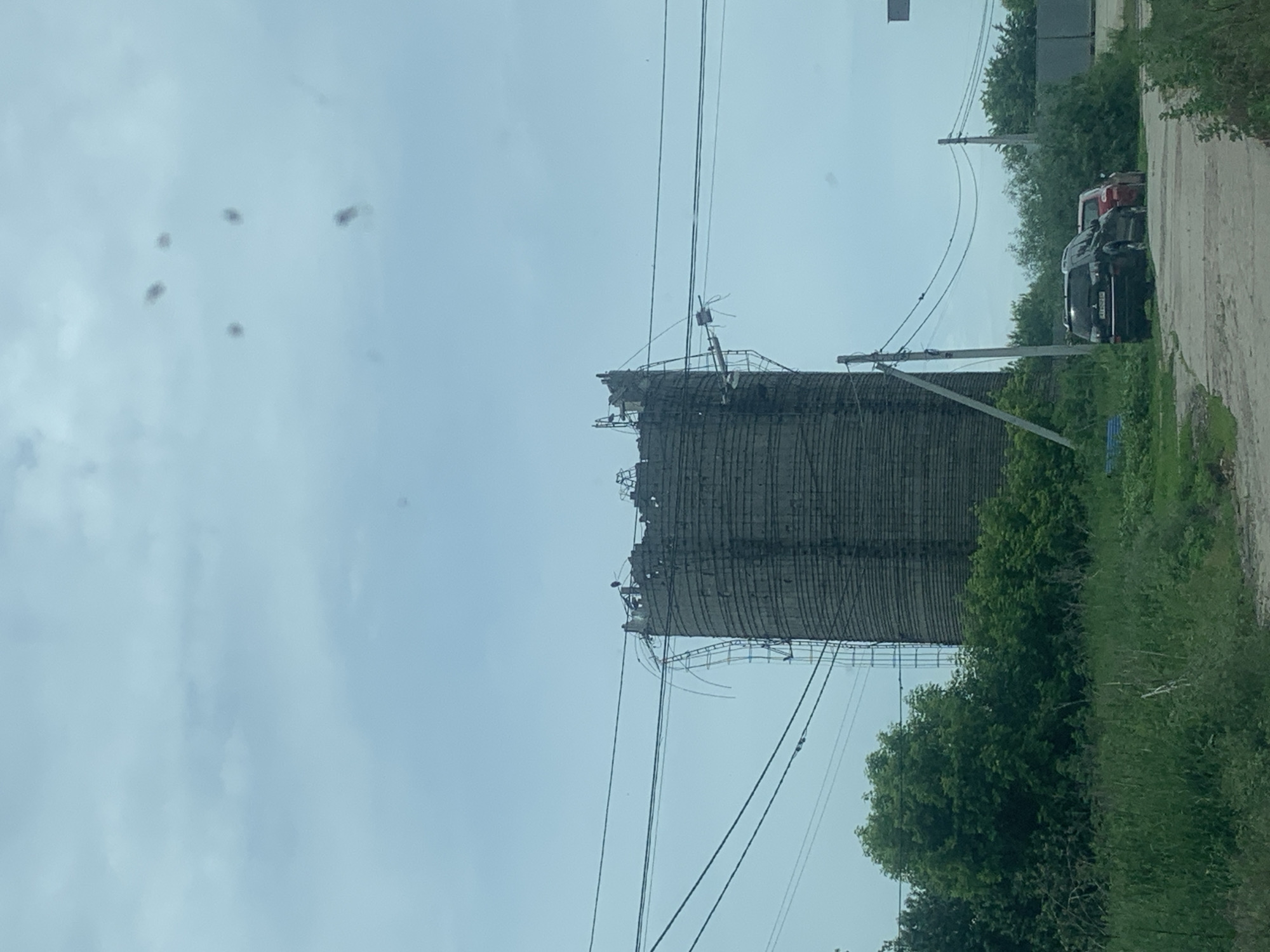 The image depicts an outdoor scene in Hostomel, Ukraine, under an overcast, light blue-grey sky. The primary subject is a tall, cylindrical industrial structure, likely a silo or tower, composed of horizontally ribbed concrete or masonry. Its upper section is heavily damaged, displaying jagged, fractured edges and exposed internal elements, including what appears to be bent rebar or structural supports. Metal ladder-like structures are attached to the exterior, particularly visible on the left side, with some sections appearing distorted or partially detached. Dark, indistinct figures, possibly birds, are perched on the damaged rim of the structure.
In the mid-ground, multiple black power lines traverse the scene, supported by several utility poles. One prominent pole stands to the right of the damaged tower, connecting to the network of wires. Below the power lines, dense green vegetation, including trees, bushes, and tall grass, covers the landscape, obscuring the base of the tower and extending into the foreground.
On the right side of the image, two vehicles are parked on a rough, unpaved or poorly maintained road/path. The closest vehicle is a dark-colored SUV, possibly a Mitsubishi Outlander, with a partially visible Ukrainian license plate beginning with "AI 90." Behind it, a red car is partially visible. The road surface exhibits cracks and uneven terrain, with grass and weeds growing along its edges. No individuals are visible in the scene. The overall environment appears to be an industrial or semi-rural area with elements of disrepair and overgrowth. Several dark, irregular smudges are present in the sky, indicative of lens debris or imperfections on the camera or window through which the photograph was taken.