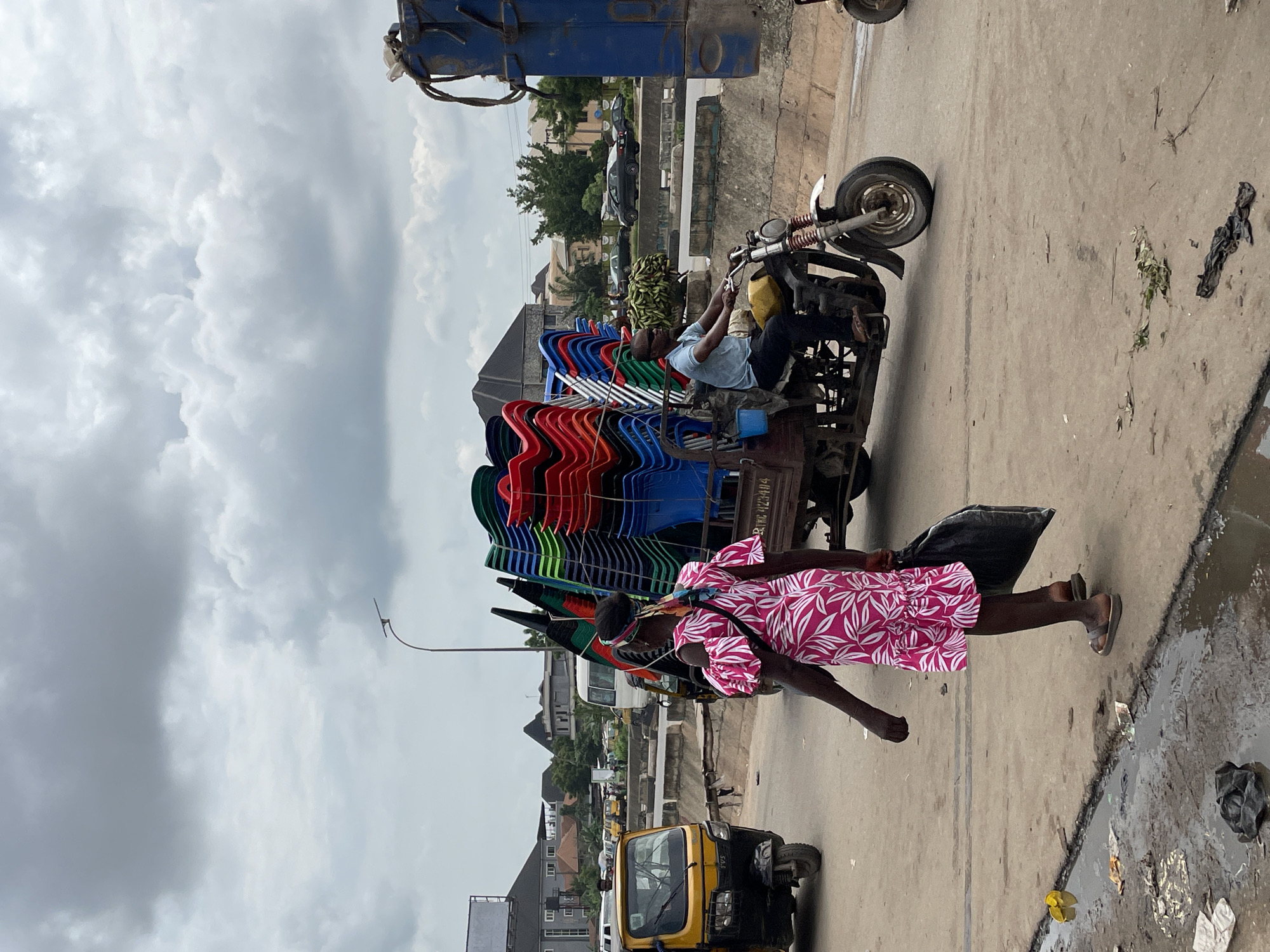 A medium shot captures a street scene in an urban environment under an overcast sky. In the foreground, a person wearing a pink and white floral dress walks away from the camera, carrying a black plastic bag. To their right, a motorized tricycle with a trailer is driven by a man wearing sunglasses and a light blue shirt. The trailer is laden with a large stack of colorful plastic chairs, predominantly red, green, and blue. The tricycle is on a paved road, with a yellow auto-rickshaw visible to the left. In the background, various buildings, trees, and other vehicles are present, including a white bus. A blue truck is partially visible on the far right of the frame. The ground is a mix of concrete and dirt, with puddles and scattered debris.