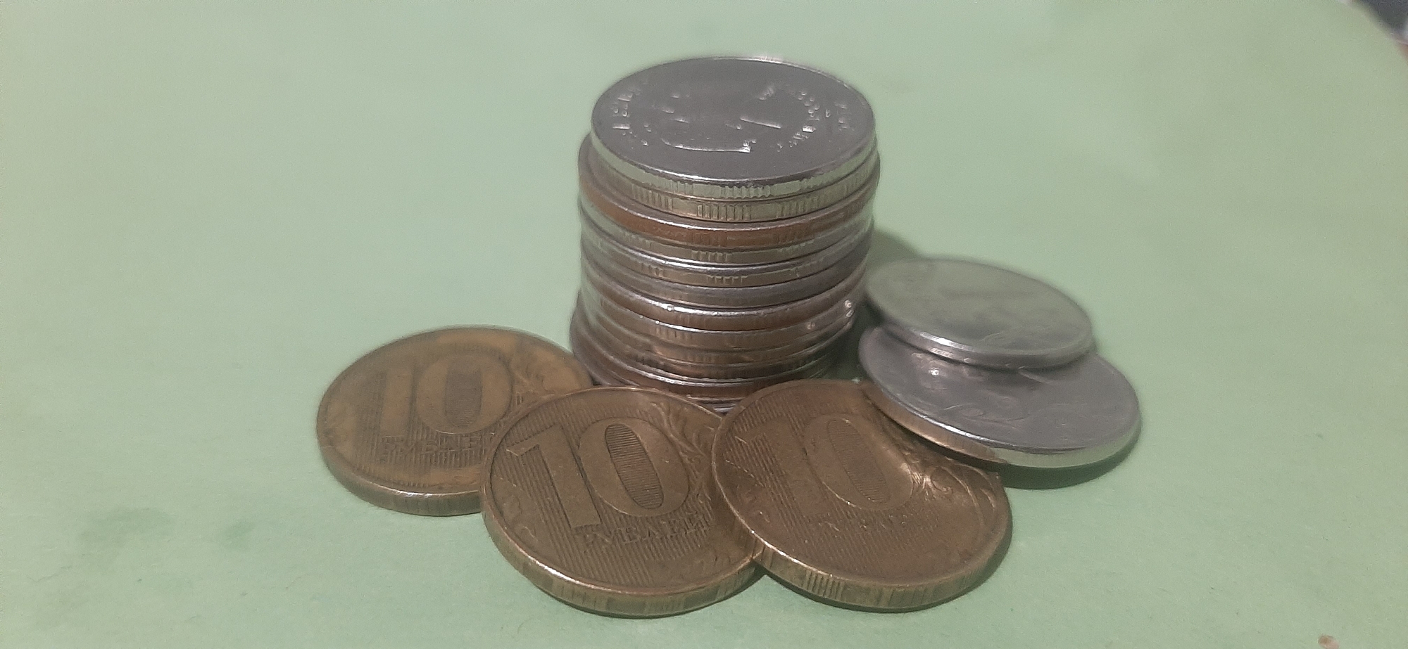 A still life image depicts a pile of coins on a light green surface. The coins are stacked in a tower in the center of the frame, with several individual coins arranged around the base. The coins appear to be of varying denominations and metals, with some exhibiting a coppery hue and others a silvery sheen. The background is a solid, pale green color. There is no indication of location in the city of Yessentukskaya, Russia.