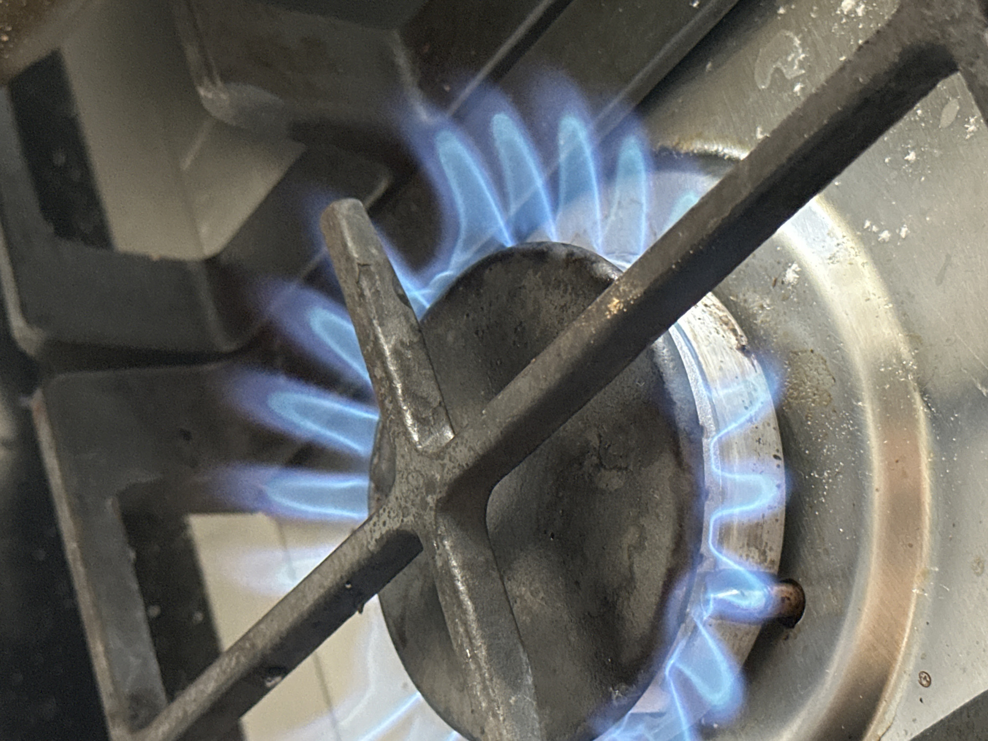A close-up, overhead view shows a gas stove burner with a blue flame igniting. The burner is made of dark metal and has a cross-shaped grate over it. The flame is bright blue and appears to be actively burning, emanating from the center of the burner. The surrounding stovetop is made of stainless steel and shows some signs of wear and residue. The background is out of focus but suggests the presence of other stove components or kitchen surfaces. The image does not provide any specific details about the city of Nevinnomyssk, Russia.