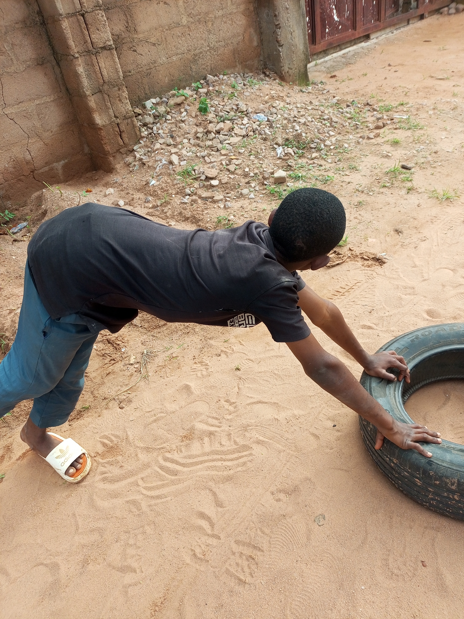 A young person with dark skin and short black hair, wearing a black t-shirt and blue pants, is on their hands and knees on sandy ground next to a worn tire. The person is reaching out to touch the tire with both hands. The environment appears to be an outdoor area with a rough, textured brown brick wall behind the person and a metal gate to the right. The ground is covered in sand with scattered small stones and some sparse vegetation. The overall impression is one of a casual or recreational activity taking place in a somewhat undeveloped urban or semi-urban setting, likely in Gombe, Nigeria, given the characteristic sandy terrain and construction style.