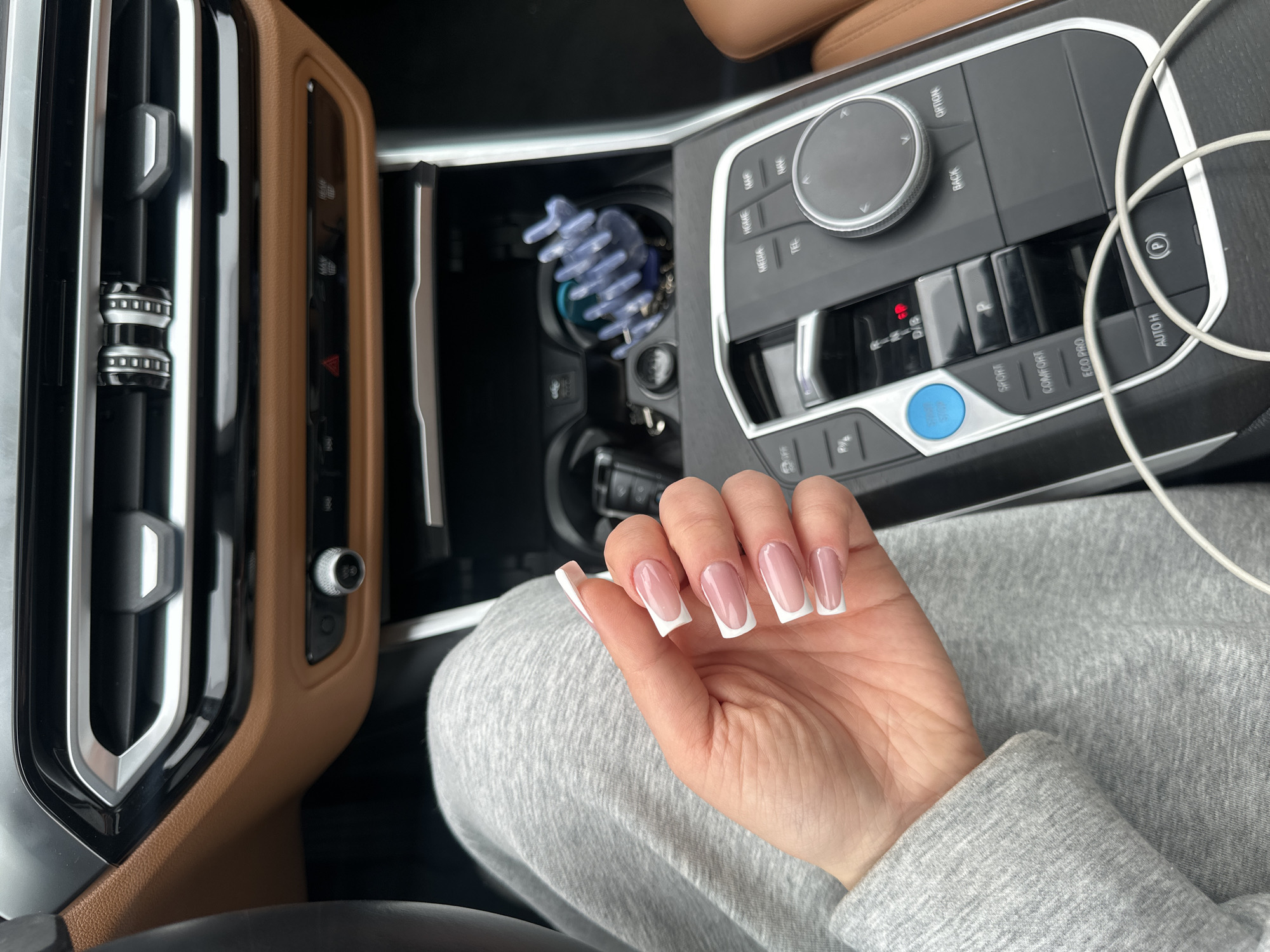 The image presents an interior view from a vehicle, specifically a modern BMW model, characterized by its brown leather dashboard trim, silver-accented air vents, and contemporary central console. The precise geographical location within Yerevan, Armenia, is not discernible from the visual information provided.
A fair-skinned human hand, featuring long, square-shaped nails with a clear pink base and prominent white French tips, occupies the left-center foreground. The hand is connected to an arm covered by a grey heathered long-sleeved garment, likely a sweatshirt or similar casual apparel, the fabric of which also covers the visible upper leg.
The central console of the vehicle displays an electronic gear selector with the "P" (Park) setting illuminated in red. Adjacent controls include a blue "START/STOP" button, driving mode selections (SPORT, COMFORT, ECO PRO), an "AUTO H" (Auto Hold) button, and an electronic parking brake. To the right of the gear selector, a rotary iDrive controller is present, surrounded by buttons for navigation, media, and telephone functions. Within the forward storage or cup holder area, a blue or purple plastic hair claw clip is visible. A white charging cable is loosely draped across the lower section of the central console. No active interaction with the vehicle controls is depicted.
