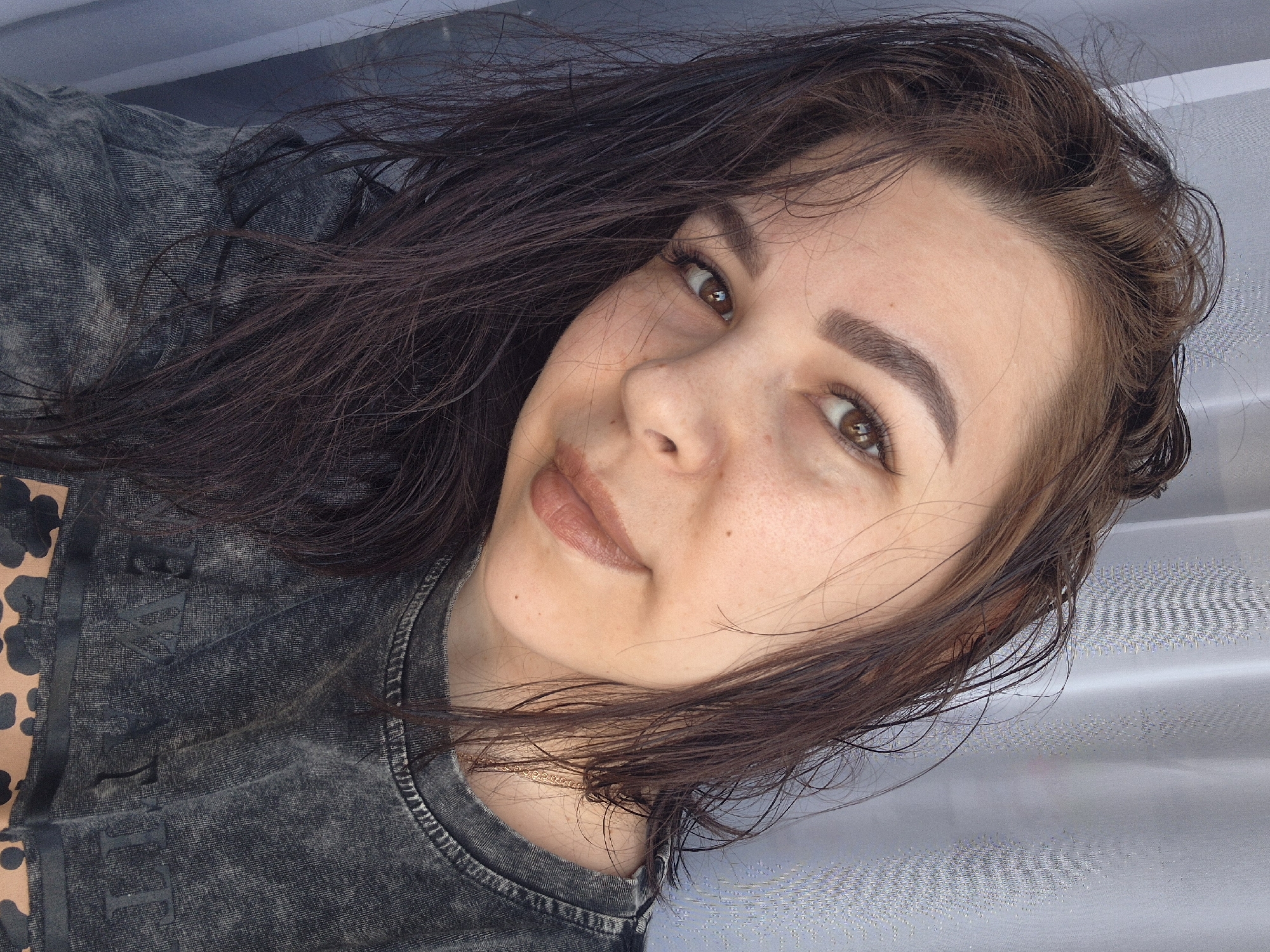 A close-up, head-and-shoulders portrait of a woman with brown hair and brown eyes. She is wearing a gray acid-wash T-shirt with dark letters on the front. The background is a textured light gray or white surface. There is nothing indicating the location is Tobolsk, Russia.