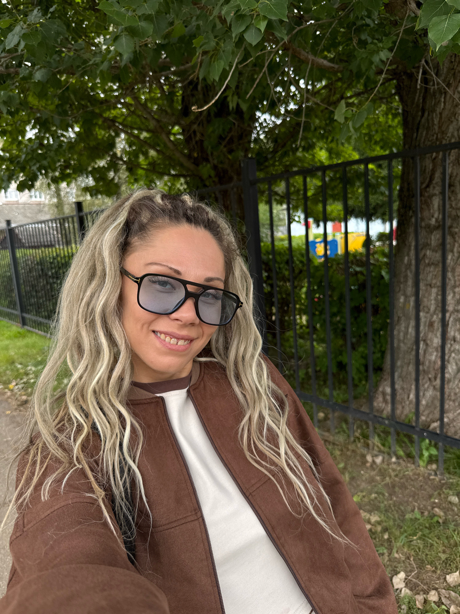 In Rusanovo, Russia, a woman with blonde, wavy hair and large, dark-rimmed sunglasses smiles at the camera. She is wearing a brown bomber jacket over a white shirt. Behind her, a black metal fence separates her from a green, leafy area with trees and some indistinct colorful objects in the background, possibly playground equipment. A building and another fence are visible to the left. The ground is covered with grass and scattered leaves.