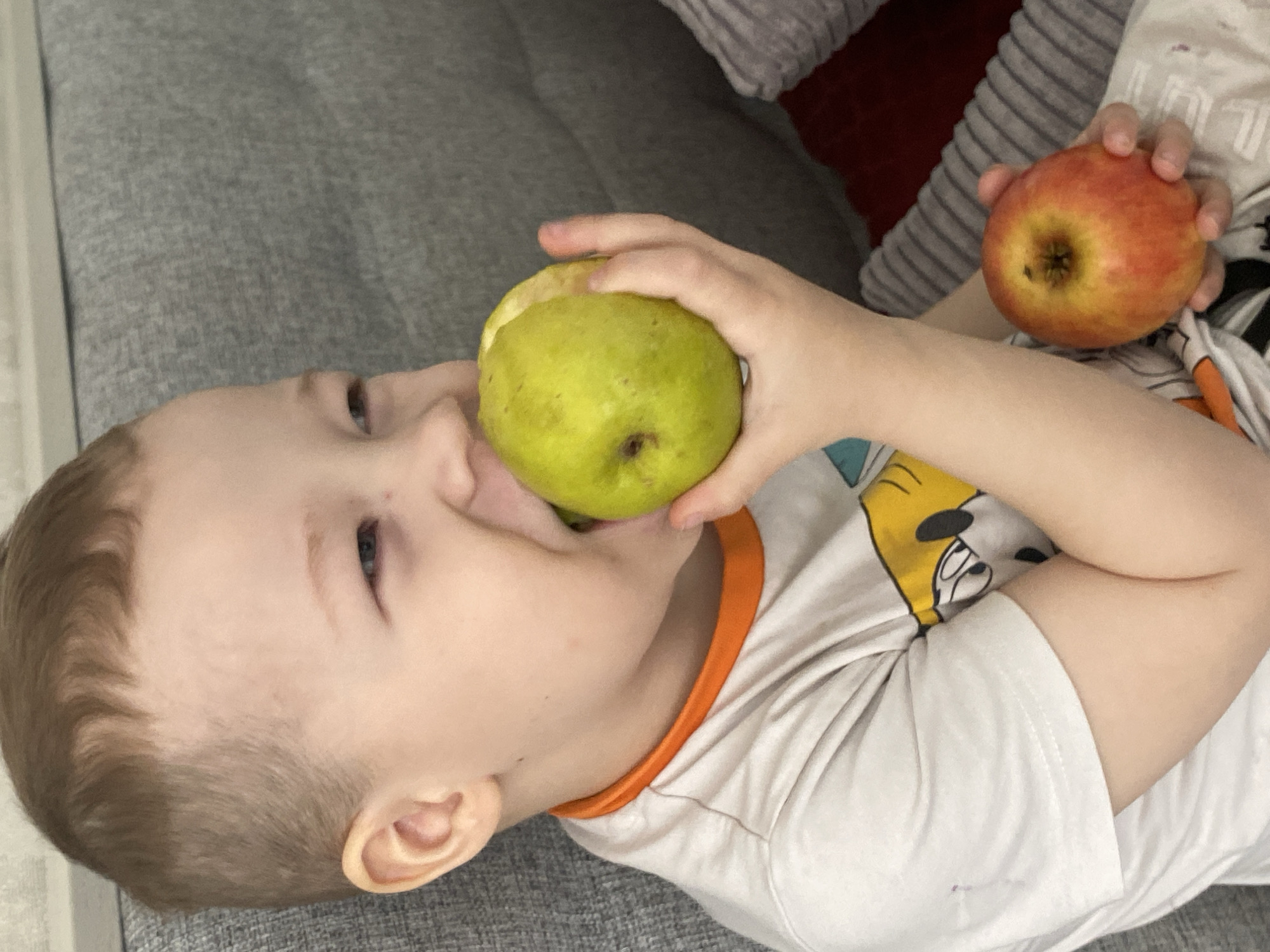 A young child is lying on a grey surface and eating a green apple. The child is holding a red apple in their other hand. The child is wearing a white t-shirt with an orange collar. The background includes a grey surface and some pillows. The location is not determinable from the image.