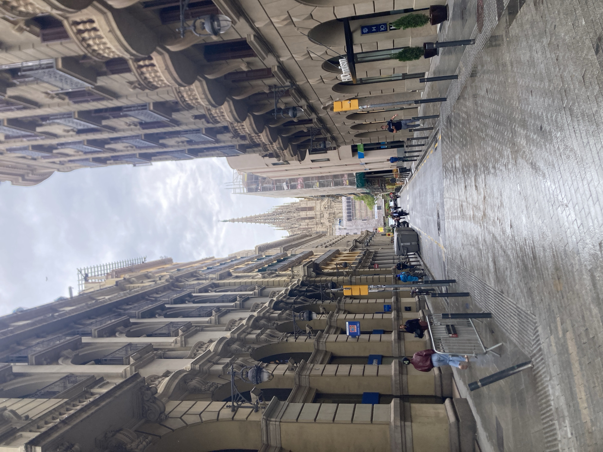 This is a wet street scene in Barcelona, Spain, featuring ornate buildings on either side of the street. The buildings have a classic architectural style with balconies and detailed stonework. A tall, cathedral-like structure is visible in the distance at the end of the street. Several pedestrians are walking along the street. The cobblestone street is wet, reflecting the surroundings. Metal barricades or barriers are set up along the sidewalks. Street lamps and hotel signs are visible. The sky is overcast.