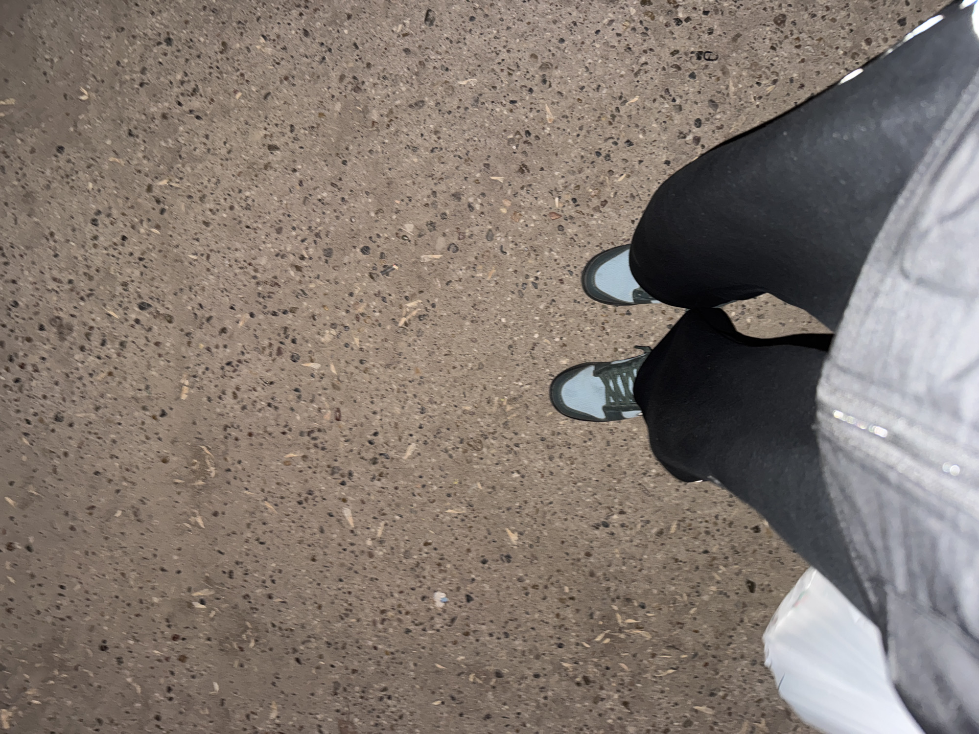 The image shows a top-down view of a person's lower body, focusing on their feet and the ground beneath. The person is wearing dark-colored pants or leggings and light blue or grey sneakers with darker laces. A portion of a light-colored outer garment, possibly a jacket or coat, is visible on the right side of the frame, along with a crumpled white plastic bag near the bottom right.
The ground is a coarse, speckled surface, resembling concrete or asphalt, with various small dark and light aggregates embedded within. The lighting is dim, suggesting it might be evening or nighttime, or a very overcast day. There are no strong shadows present. The overall impression is an outdoor setting, likely a sidewalk or paved area in an urban or suburban environment. No specific activity beyond standing or pausing is clearly depicted. There is no visible text in the image. The location is Kazan, Russia, though no specific visual cues within the image confirm this.