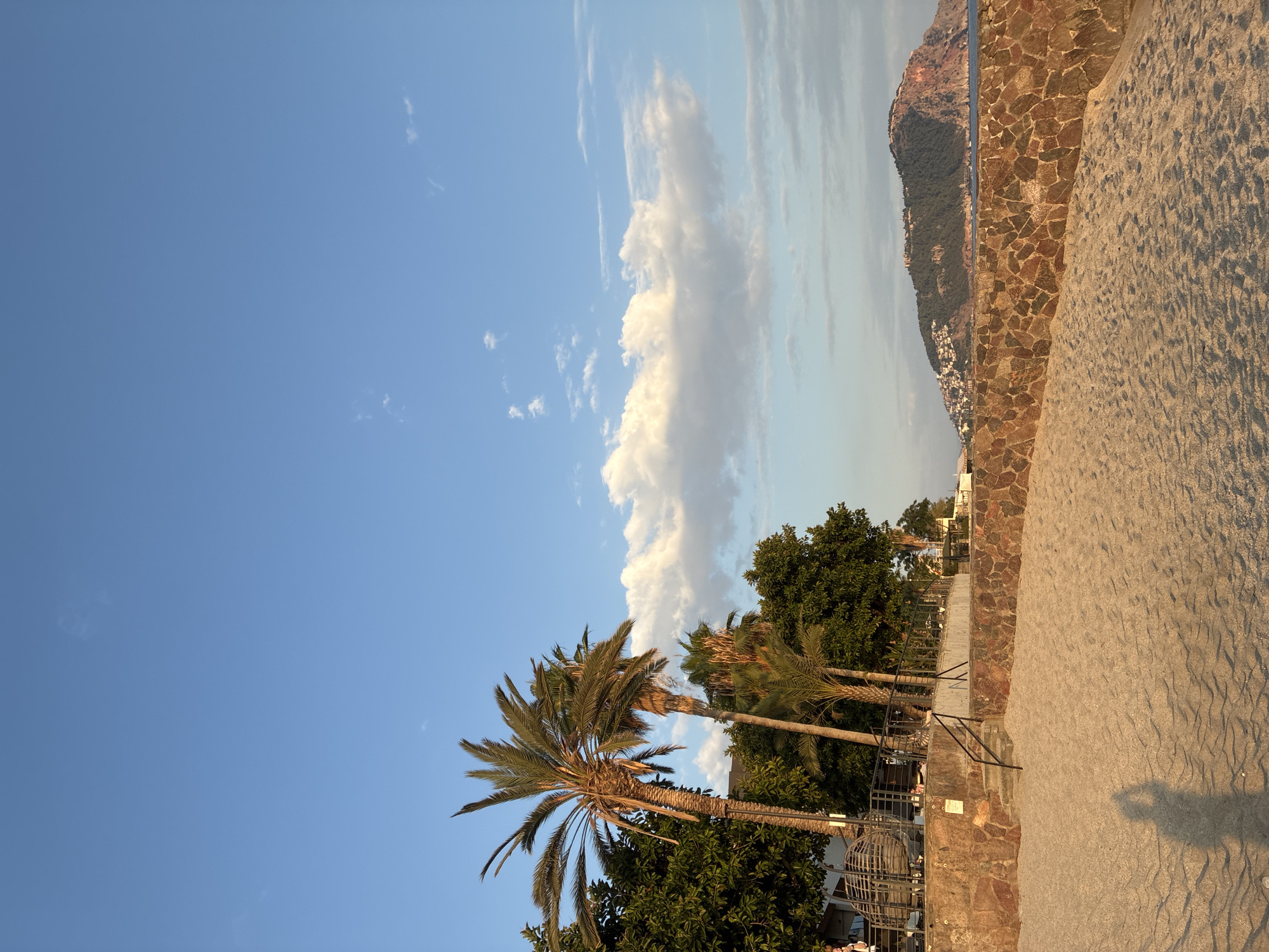 This image depicts a tranquil beach scene in Alanya, Turkey, likely during the late afternoon or early evening based on the golden light illuminating the sand and the shadows. In the foreground, soft, pale sand stretches across the frame, marked by the distinct shadow of a person holding a camera, indicating the vantage point of the photographer.
Behind the sand, a low, rugged stone wall separates the beach from a slightly elevated area. This area features lush green trees and several palm trees, some with dry fronds, interspersed with buildings that appear to be part of a resort or restaurant complex, suggested by the presence of outdoor seating and railings. A set of concrete stairs with black metal railings leads up from the sand to this elevated section.
In the distance, a prominent, verdant hill rises from the sea, dotted with white buildings and topped with structures that suggest a historic castle or citadel, a characteristic feature of Alanya's skyline. The sea is visible beyond the stone wall, meeting the horizon under a clear blue sky punctuated by soft, wispy clouds. The overall atmosphere is one of peace and natural beauty, characteristic of a Mediterranean coastal resort town.