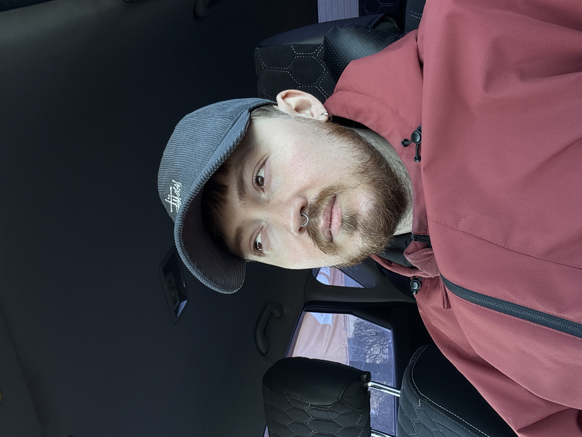 A man is sitting in what appears to be the driver's seat of a car. He is wearing a gray Stüssy cap and a maroon jacket. The interior of the car is visible, including the seat and door. Outside the window, there are trees and what appears to be a building. The photo was taken in Chaikovsky, Russia.