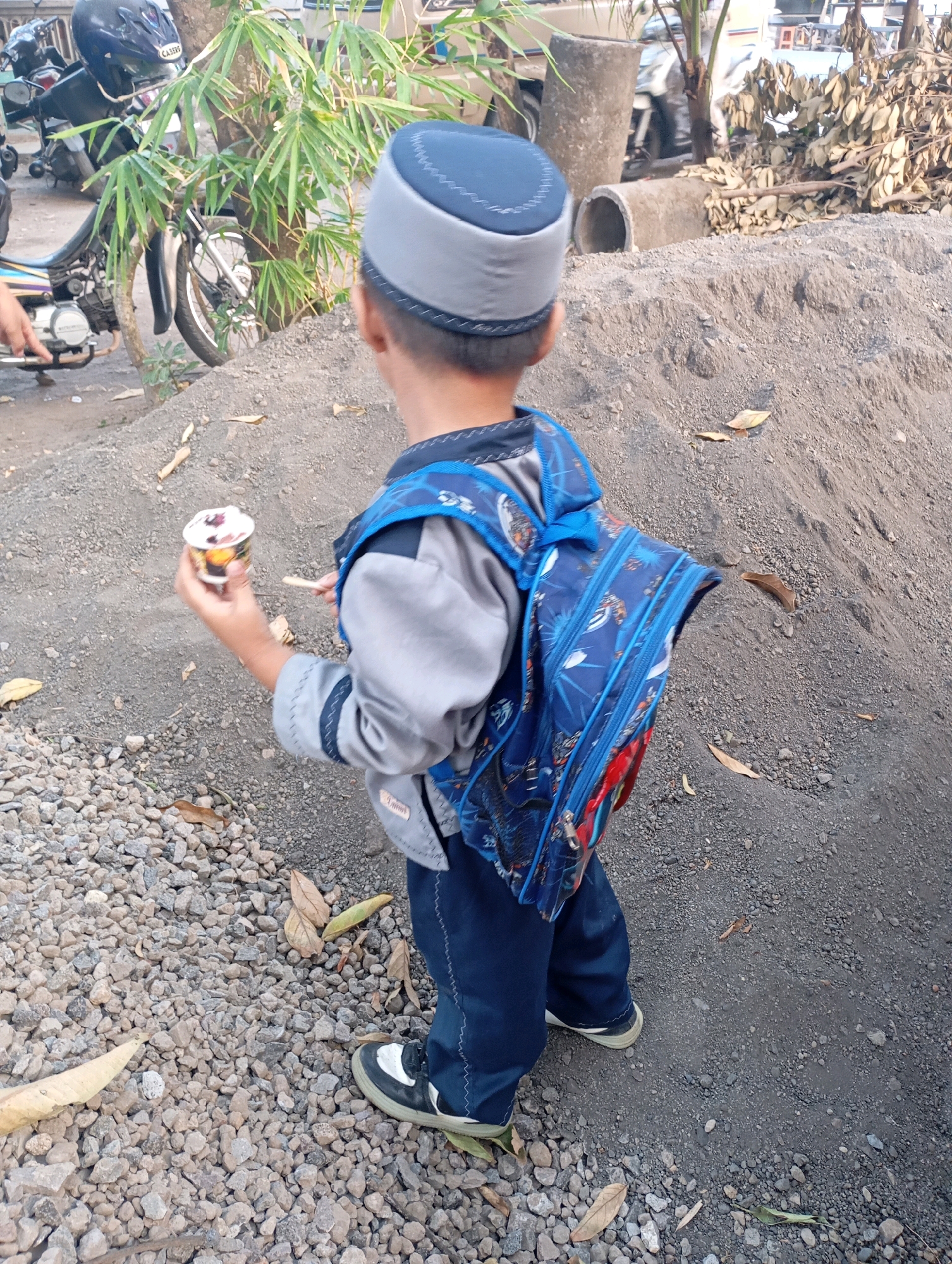 In Ketegan, Indonesia, a young boy is pictured from behind, wearing a gray and blue peci cap and a blue backpack with a graphic print. He is holding a small cup of ice cream in his left hand and a spoon in his right. The boy is standing on a surface composed of small gray pebbles and dirt, with a large pile of sand to his right. In the background, a motorcycle is parked near a tree with green foliage, and several concrete pipes and fallen leaves are visible. The overall environment suggests an outdoor, possibly construction or informal roadside setting.