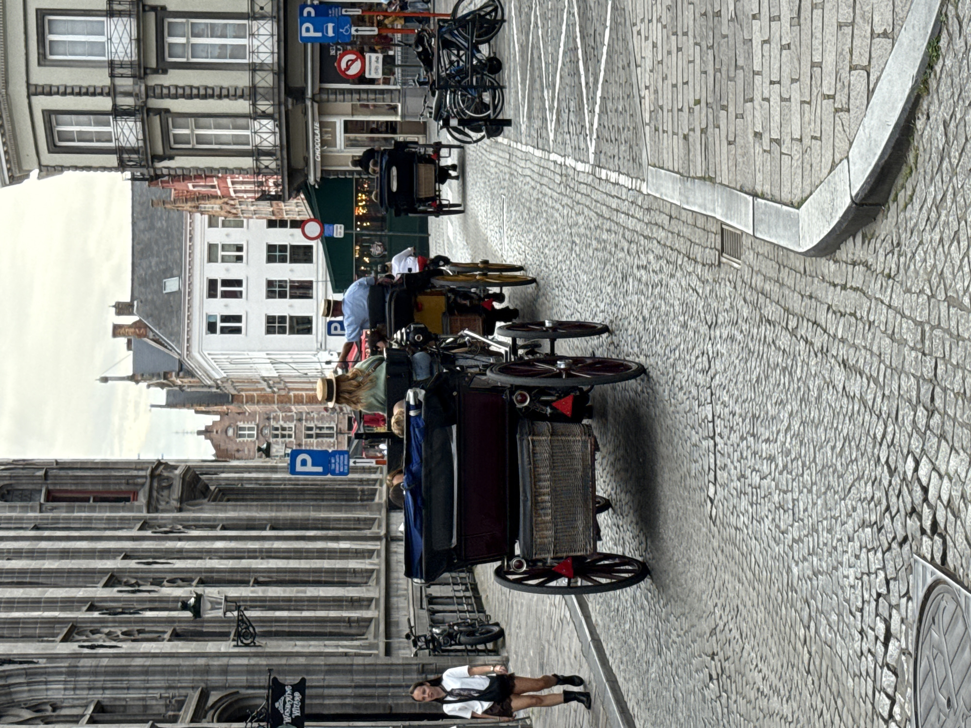The image depicts a street scene in the historic city of Bruges, Belgium, characterized by cobblestone streets and traditional European architecture. In the foreground, a horse-drawn carriage is prominently featured, with its dark red body and wicker basket. A woman in a white top and brown shorts is walking on the left side of the street, looking towards the viewer. Behind her, a black motorcycle is parked.
Further down the street, two more horse-drawn carriages are visible. The first carriage has a yellow frame and is carrying passengers, including a person wearing a blue shirt and a hat. The second carriage, further in the distance, is dark and also appears to be carrying passengers. The buildings lining the street showcase classic architectural styles, with stone facades and numerous windows. One building on the right displays a sign that reads "CHOCOLATE."
Several parked bicycles are visible on the right side of the street, and blue parking signs with a "P" are posted on poles. The sky is overcast, suggesting a cloudy day. The overall atmosphere of the scene is one of historical charm and leisurely tourism, typical of Bruges.