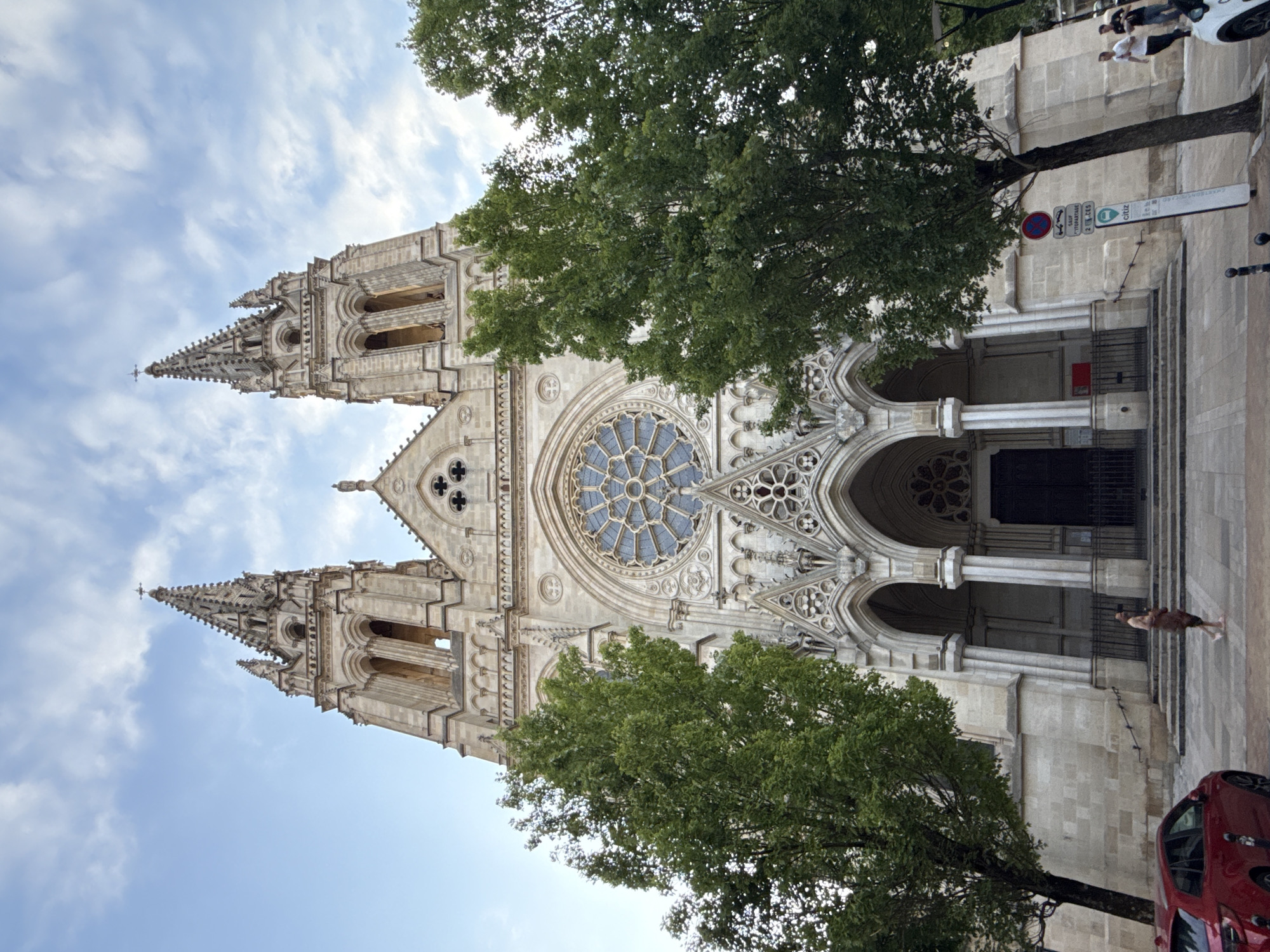 The image captures a low-angle view of the Cathédrale Saint-André de Bordeaux, a Gothic cathedral in Bordeaux, France. The cathedral's facade is dominated by two prominent spires, a large rose window, and intricate architectural details. Two large trees flank the entrance, partially obscuring the building. In the foreground, a red car is parked on the left, and on the right, a signpost and some pedestrians are visible. One person, a woman, is walking towards the cathedral entrance, carrying a bag. Two men are walking away from the camera on the far right. The sky above is partly cloudy, suggesting it is daytime.