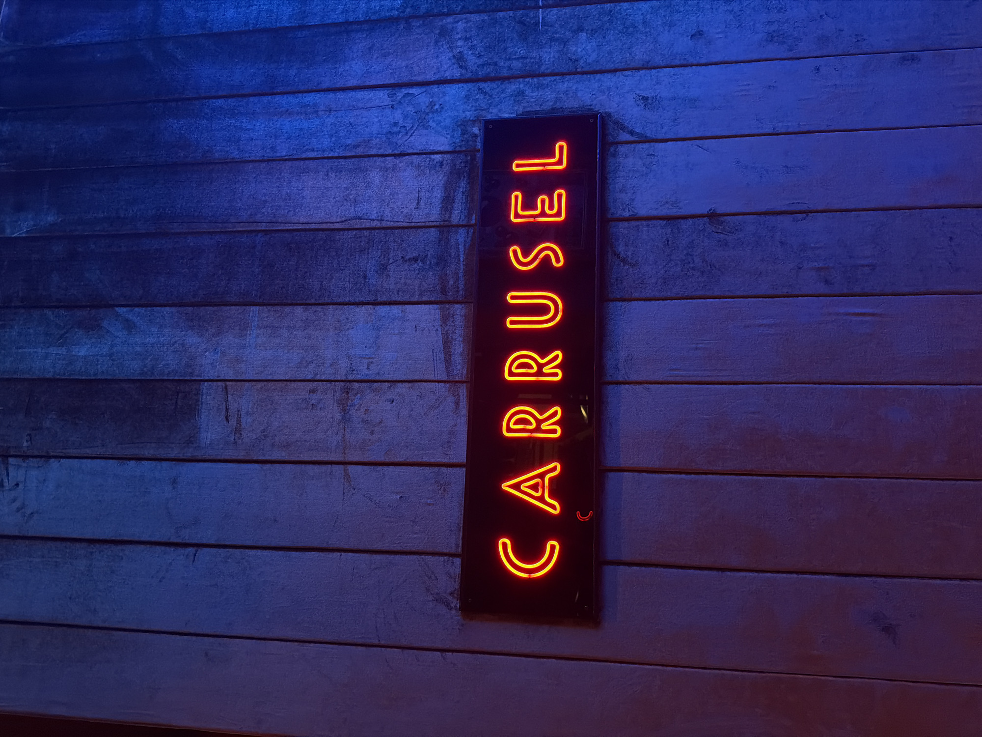 A vertical black sign with the word "CARROUSEL" in orange neon lettering is mounted on a wooden wall. The wall is made of horizontal planks with a blue and grey hue. The photograph appears to have been taken in low light conditions.