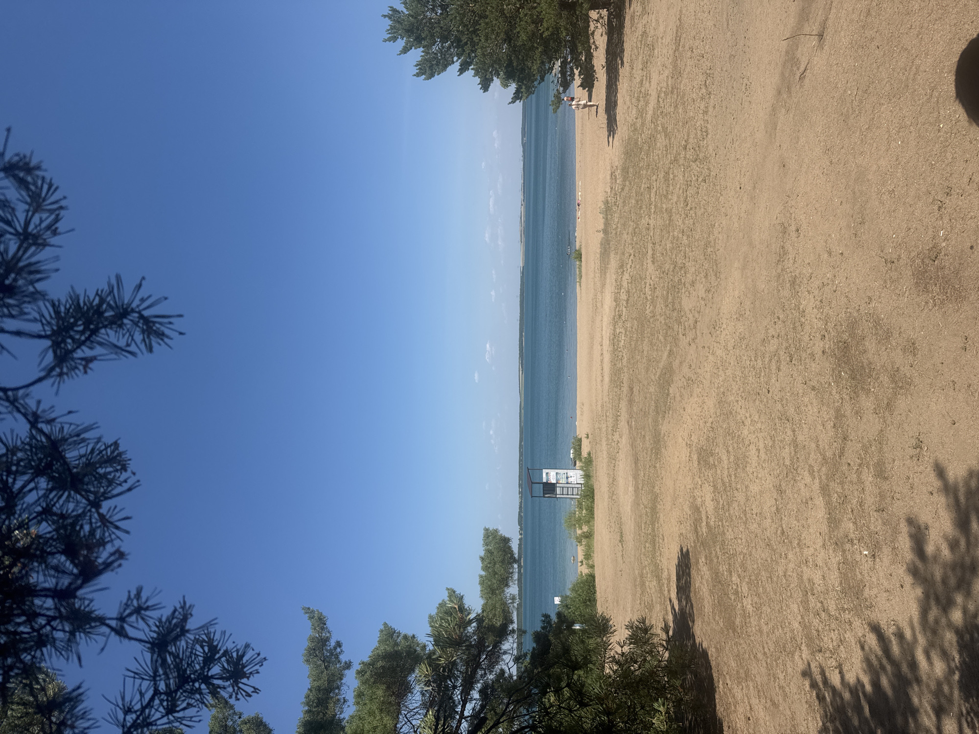This image depicts a bright, sunny day at a sandy beach with a clear blue sky. In the foreground, the dark silhouettes of evergreen trees frame the left side of the scene. The beach itself is composed of light-colored sand, with sparse patches of dry grass. A calm, blue body of water stretches across the middle ground, meeting the horizon under the expansive sky.
Several objects and subtle details are visible. A white lifeguard tower stands near the water's edge, suggesting a recreational area. In the distance, a few people can be seen on the beach and possibly in the water, indicating a relaxed atmosphere. There is also a shadow of a person cast on the sand, implying the time of day is likely midday or afternoon due to the strong sunlight. The overall impression is one of a peaceful summer day in a natural setting, possibly a lake or a very calm sea. The location context suggests this is Antonovka, a rural district in Kazakhstan. No visible text is present in the image.