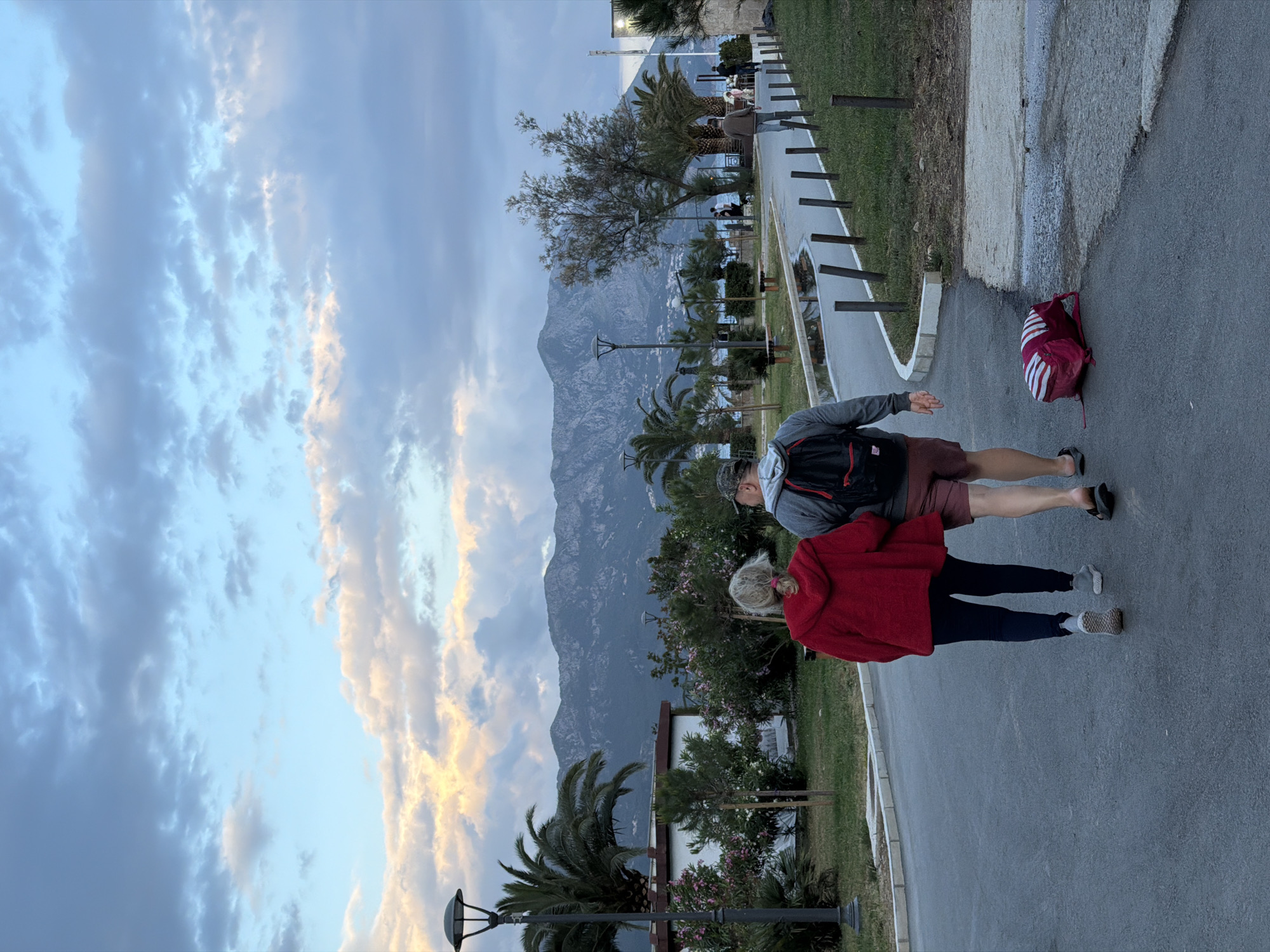 The image shows a couple walking on a paved path in Budva, Montenegro, with a mountain range visible in the background. The sky is cloudy with patches of blue. Palm trees and various plants line the path. A red and white backpack is visible lying on the pavement. The man is wearing a gray shirt, red shorts, and a backpack. The woman is wearing a red sweater and dark leggings.
