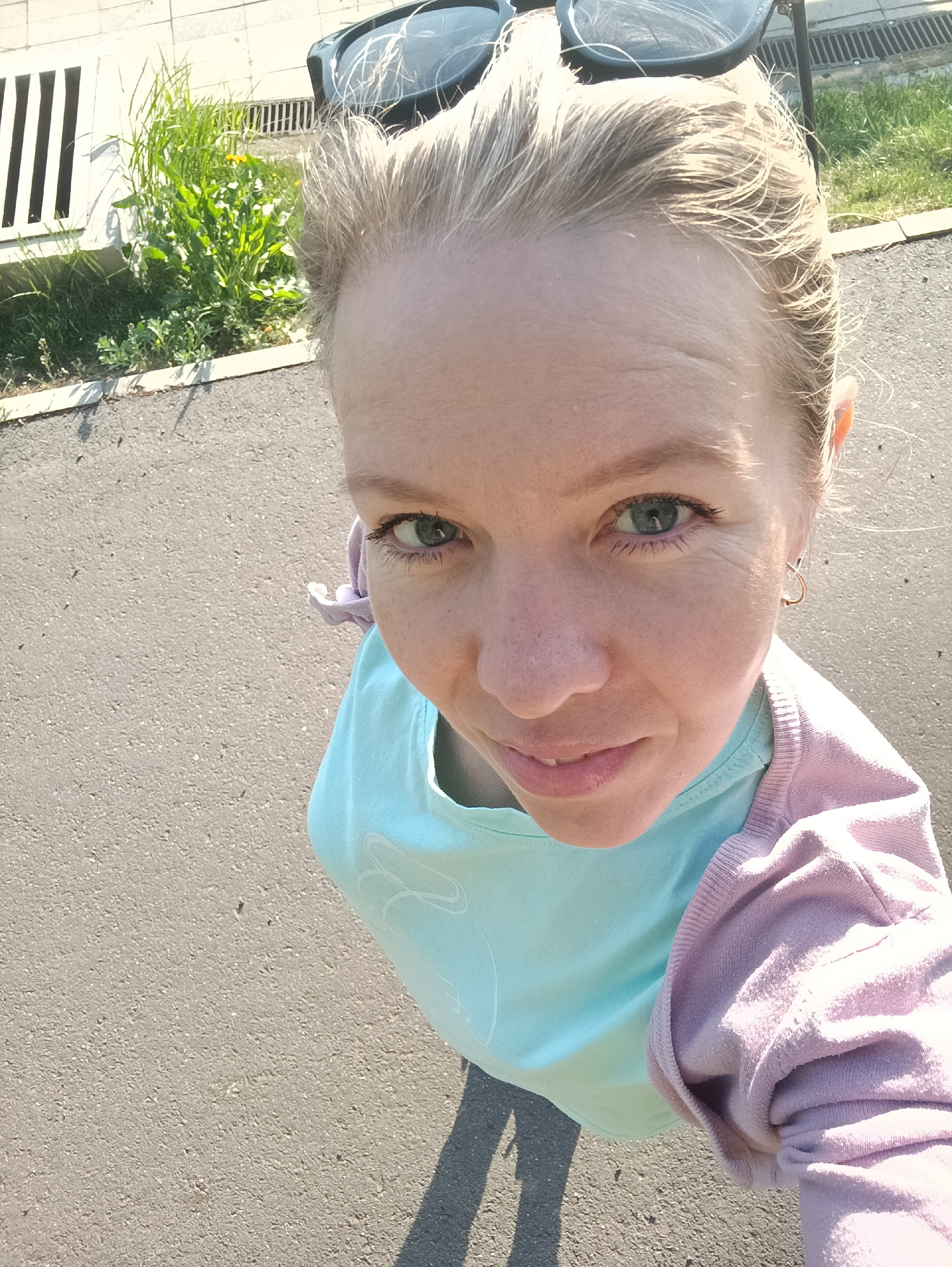 A woman is taking a selfie while standing on an asphalt surface, likely a street or path, in Chaikovsky, Russia. She has blonde hair, fair skin, and is wearing a light blue t-shirt with a light purple sweater. Sunglasses are resting on her head. In the background is a curb, grass, and a drainage grate. The sunlight indicates it's likely daytime.