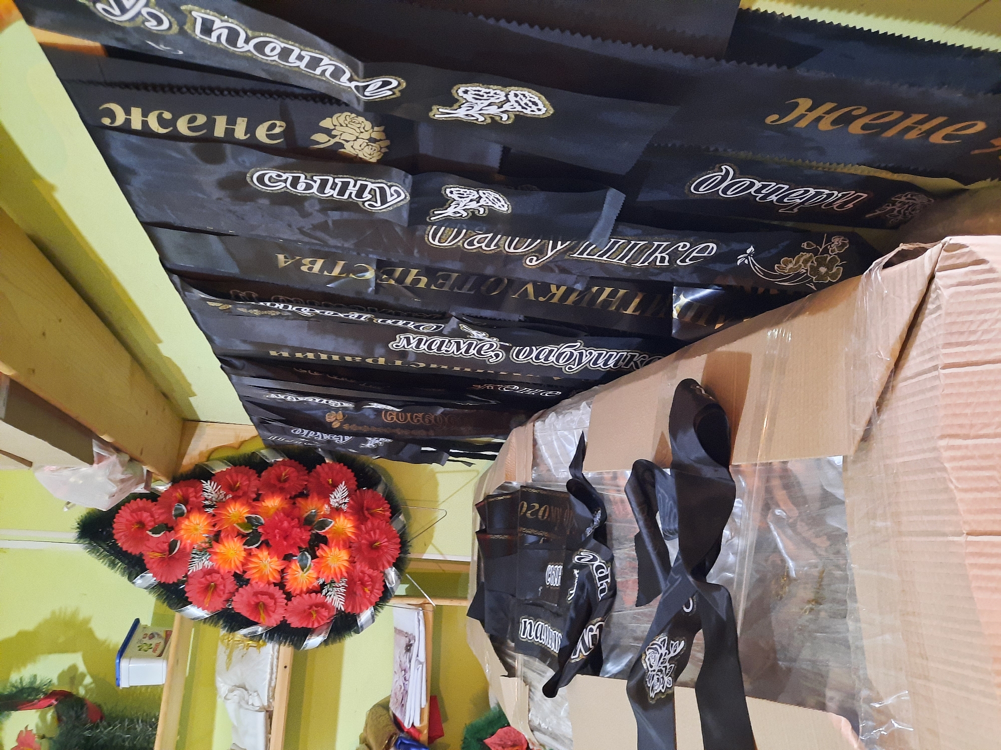 The image shows an interior space with various items related to funeral services. Several black ribbons with gold lettering and floral designs hang from the ceiling, with phrases like "жене" (wife), "сыну" (son), "дочери" (daughter), and "маме, бабушке" (to mother, grandmother) in Russian. A floral wreath made of red and orange flowers is visible in the lower left corner. A cardboard box is positioned to the right, with more ribbons draped over it. Some writing on the ribbons is partially obscured. The environment appears to be a storage or work area within a shop or facility. The scene is situated in Krasnogorskoye, Russia.