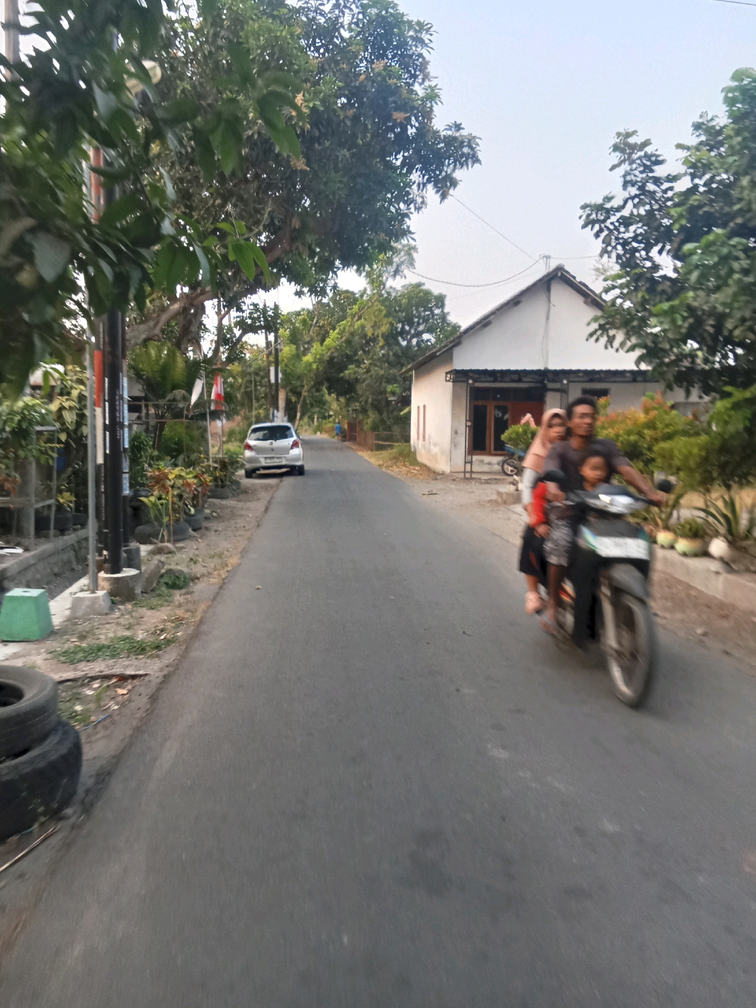 The image depicts a residential street scene in Jombang, Indonesia, likely in the early morning or late afternoon given the lighting. A paved road runs through the center of the frame, flanked by houses and vegetation.
In the foreground, to the left of the road, are stacked tires and some greenery. Further down the road, a silver car is parked on the side. A prominent feature is a motorcycle traveling towards the viewer, carrying three people. The driver, a man, is in the front, followed by a woman wearing a hijab and a child in the middle. Another child is seated in front of the driver. The motorcycle is in motion, indicated by motion blur.
On the right side of the road, a single-story house with a white facade and a dark tiled roof is visible, partially obscured by trees. There are potted plants and small trees in front of the house. A power pole with wires runs along the left side of the road. The overall environment appears to be a quiet, rural or suburban area with a mix of residential housing and natural foliage.