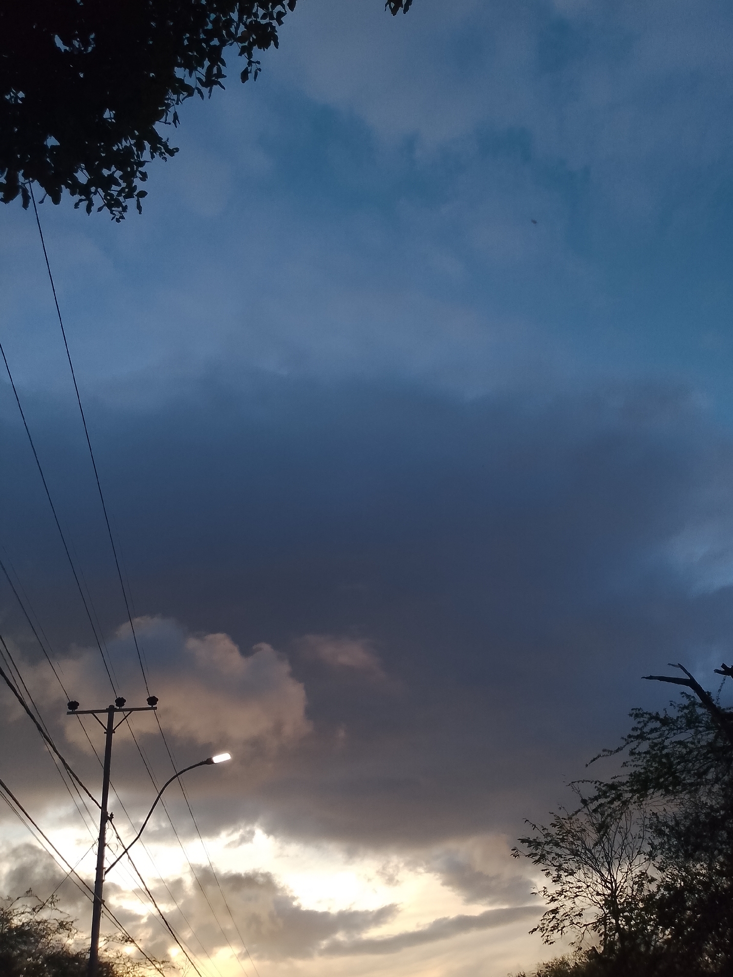 The image depicts a sky at dusk with dramatic clouds. The upper part of the frame is dominated by a dark, heavy cloud mass, transitioning to lighter shades towards the horizon. A few scattered, lighter clouds are visible near the bottom. In the lower left quadrant, a telephone pole with multiple wires and a lit street lamp stands prominently. Silhouetted trees frame the bottom right corner of the image. The scene appears to be set in an outdoor urban or semi-urban environment. The image location is identified as Santa Ana, Venezuela.