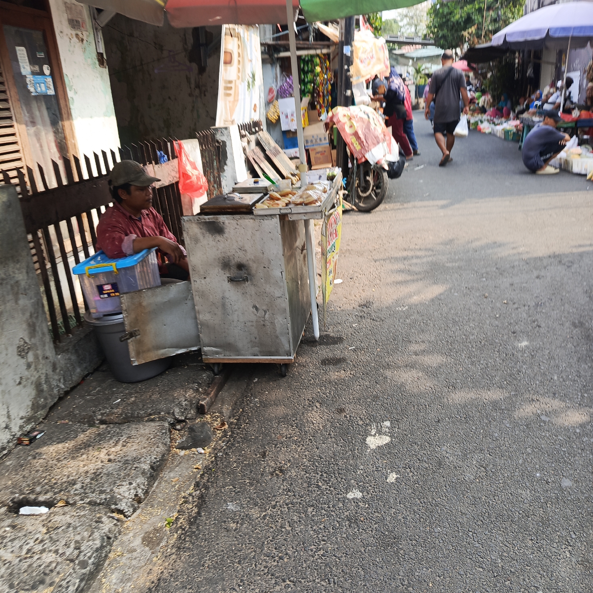 A street scene in South Jakarta, Indonesia, depicts a vendor operating a food cart on a paved road adjacent to a building. The vendor, a person wearing a red shirt and a cap, is seated behind the cart, which is equipped with a griddle and trays of food items. A blue plastic container is placed on top of a larger grey container next to the vendor. The street is lined with various stalls and shelters, suggesting a market or roadside commercial area. Several people are visible in the background, some walking and others engaged in activities related to the market. The overall environment suggests a bustling, outdoor commercial setting during daylight hours.