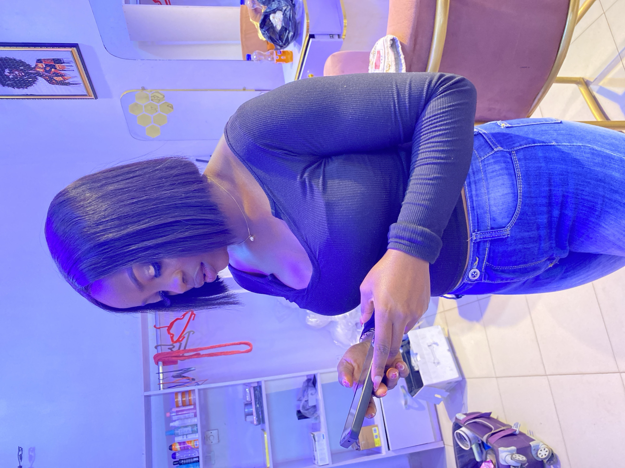 A young woman is depicted indoors in what appears to be a commercial establishment, possibly a beauty salon or boutique, under strong blue and purple artificial lighting. The woman, with short, dark, straight hair, is leaning forward and looking down at a black smartphone she holds in both hands. She is wearing a long-sleeved black ribbed top, blue denim jeans, and a light-colored necklace with a small pendant. Her fingernails are painted pink or red.
The background features a light-colored wall adorned with a framed picture showcasing a colorful, stylized figure, and a decorative piece composed of several golden hexagonal shapes. Below these, white shelves display numerous bottles and containers, suggesting beauty or cosmetic products, along with some red clothes hangers. To the right, a plush pink armchair with a golden frame is partially visible, draped with a white towel. The floor is tiled in white. A purple wheeled trolley or bag, containing various items, sits on the floor. Other incidental objects include plastic bottles and a black plastic bag. The woman's expression is neutral and focused on her activity with the phone. No discernible text is visible on any objects. The overall ambiance is cool and illuminated by the prominent blue light.