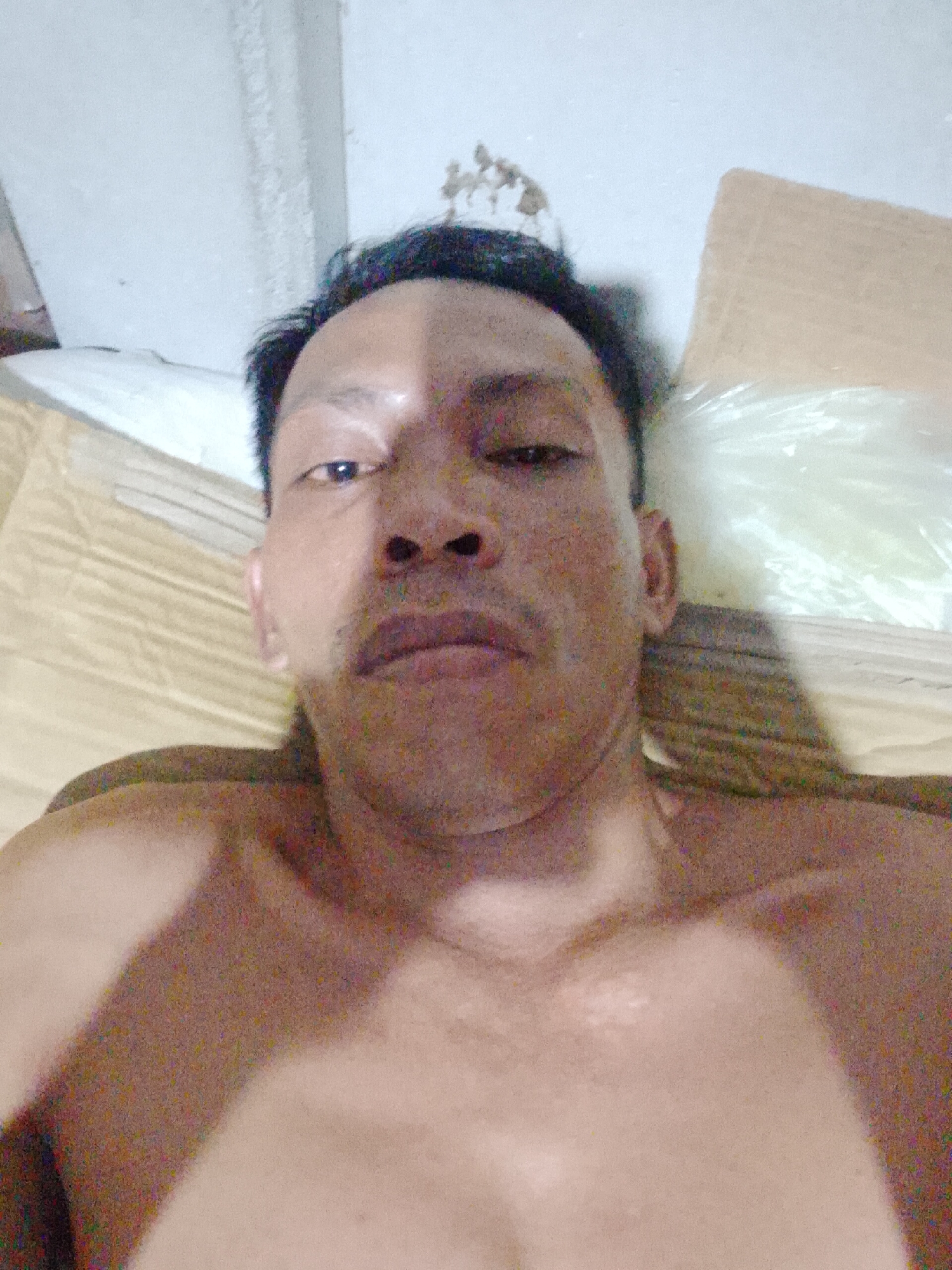 The image is a selfie of a shirtless Asian man. He is lying down, possibly in a bed. There is a cream-colored sheet beneath him. In the background are sections of a white ceiling and a cardboard box covered in plastic. The location is suggested as Bukit Ketok, Indonesia. There are no other visible subjects, objects, actions, or interactions.