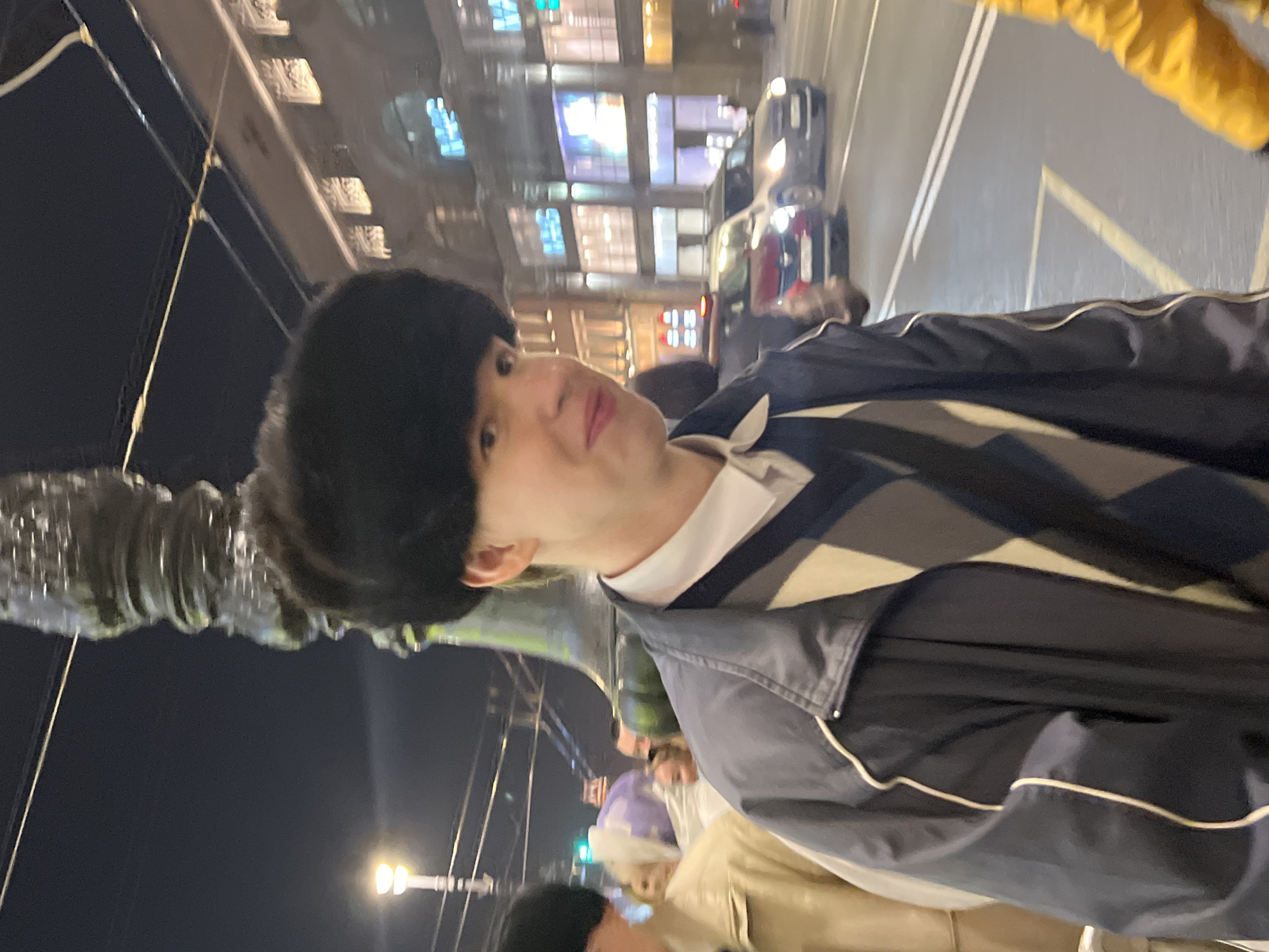 A male person wearing a dark fur hat and a patterned sweater under a dark jacket is partially visible in the foreground, possibly on a sidewalk. To the left of the person is a decorative metal structure, and behind the person is a nighttime urban scene with buildings illuminated by streetlights and signs. Cars are visible on the street, moving in the direction consistent with traffic flow. The location is likely the city of Saint Petersburg, Russia. Other people are partially visible in the background, indicating a populated area.