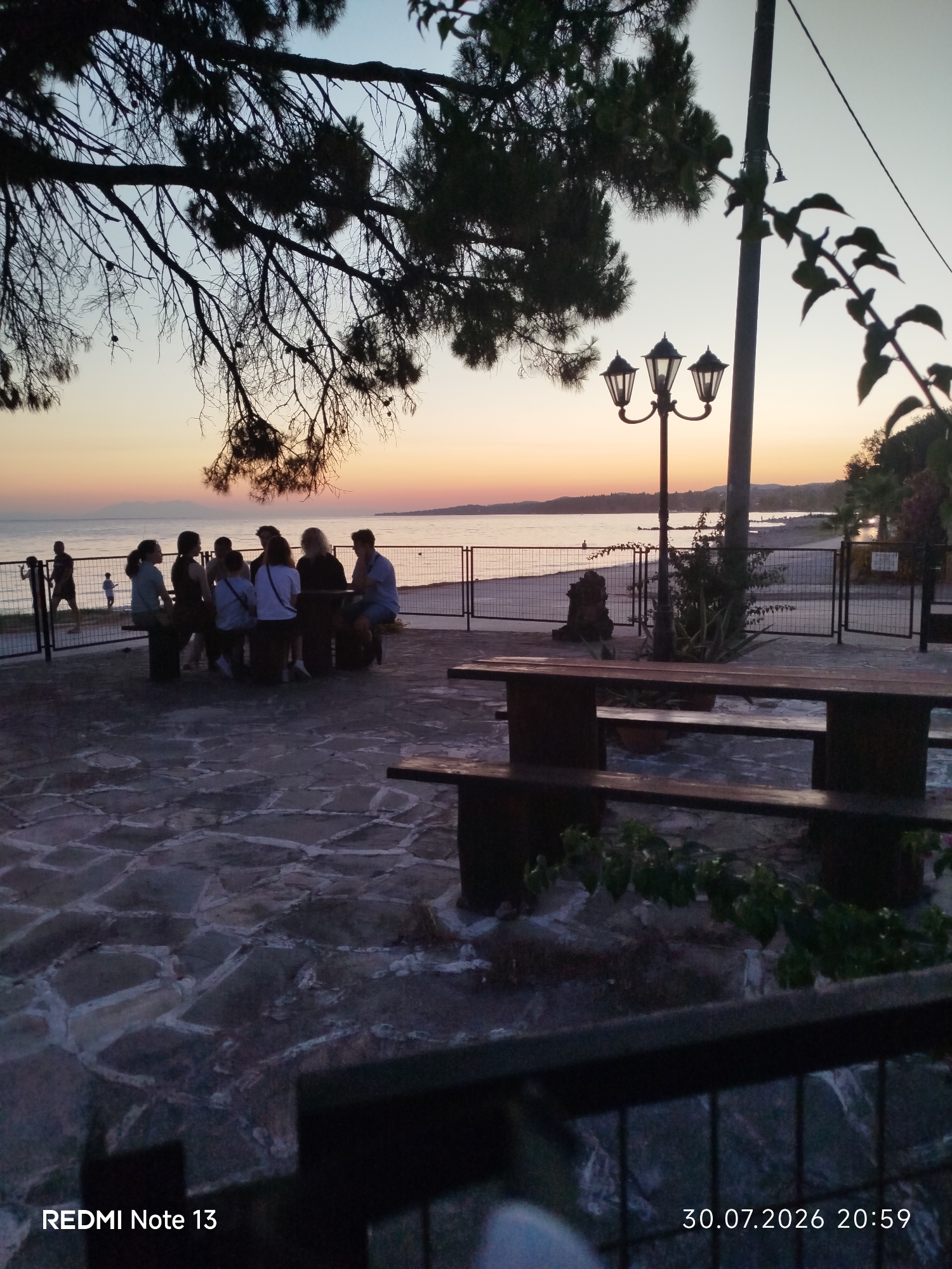 The image depicts a tranquil scene at dusk, likely in Sithonia Municipal Unit, Greece, by the sea. In the foreground, a stone-paved area features wooden picnic tables and benches, suggesting an outdoor dining or gathering space. A dark metal fence separates this area from the beach. Beyond the fence, a sandy beach stretches along the calm sea, with a faint horizon visible in the distance. A lamppost with three globes stands near the fence, illuminating the evening.
Several people are gathered on benches near the fence, some appearing to be conversing. To the left, two silhouettes of individuals are standing, and a small child is visible on the beach. The sky is painted with soft hues of orange and pink, indicating the setting sun. Overhanging tree branches frame the top of the image, adding a natural element to the composition. The overall atmosphere is peaceful and serene, characteristic of a seaside location at twilight.