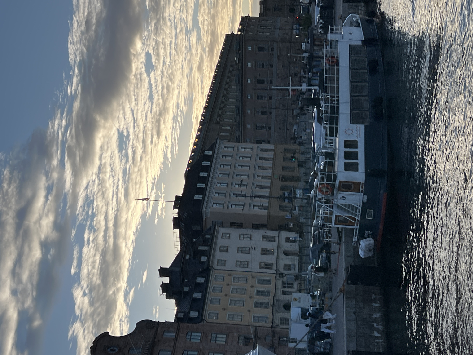 The image depicts a waterfront scene in Stockholm, Sweden, likely in the Gamla Stan district, given the historical architecture. In the foreground, a dark-hulled passenger boat with white upper decks is docked at a stone quay. The boat has several rectangular windows along its side and a visible life preserver. Behind the boat, a line of historic buildings with multiple stories and gabled roofs is visible, bathed in the soft light of what appears to be late afternoon or early morning. The sky above is a mix of blue and scattered, illuminated clouds, suggesting dynamic weather conditions. People are seen walking along the quay near parked cars and a white utility vehicle. The water in the foreground is choppy, reflecting the light and the sky. In the background, a large, grand building with a long facade and numerous windows dominates the skyline, possibly a palace or a significant governmental structure. A flag flies from a pole on this building. The overall atmosphere is one of urban activity against a backdrop of historic architecture and natural light.