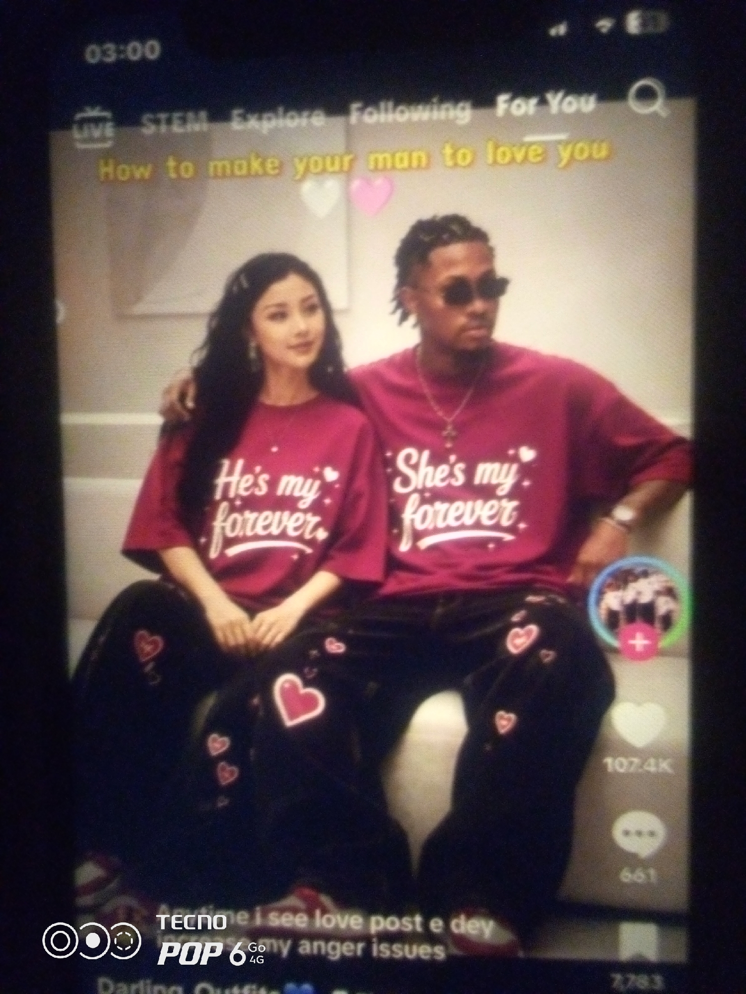The image depicts a screenshot of a video call or social media platform, featuring a young couple. The man and woman are both wearing matching maroon t-shirts with white text. The woman's shirt reads "He's my forever" and the man's shirt reads "She's my forever." Both are also wearing dark pants adorned with heart patterns. The background appears to be a brightly lit indoor setting, possibly a living room, with a couch visible. The screen displays various interface elements, including a timestamp "03:00", "LIVE", "STEM Explore Following For You", a search icon, and text overlays that read "How to make your man to love you" and "Anytime i see love post e dey Darling Outfits". There are also social media interaction icons such as likes (107.4K) and comments (661). The presence of "TECNO POP 6 Go 4G" at the bottom suggests the video is being viewed on a Tecno smartphone. The context of the video and the text suggest a focus on relationships and love. There is no indication of any specific location within Howayi, Nigeria.