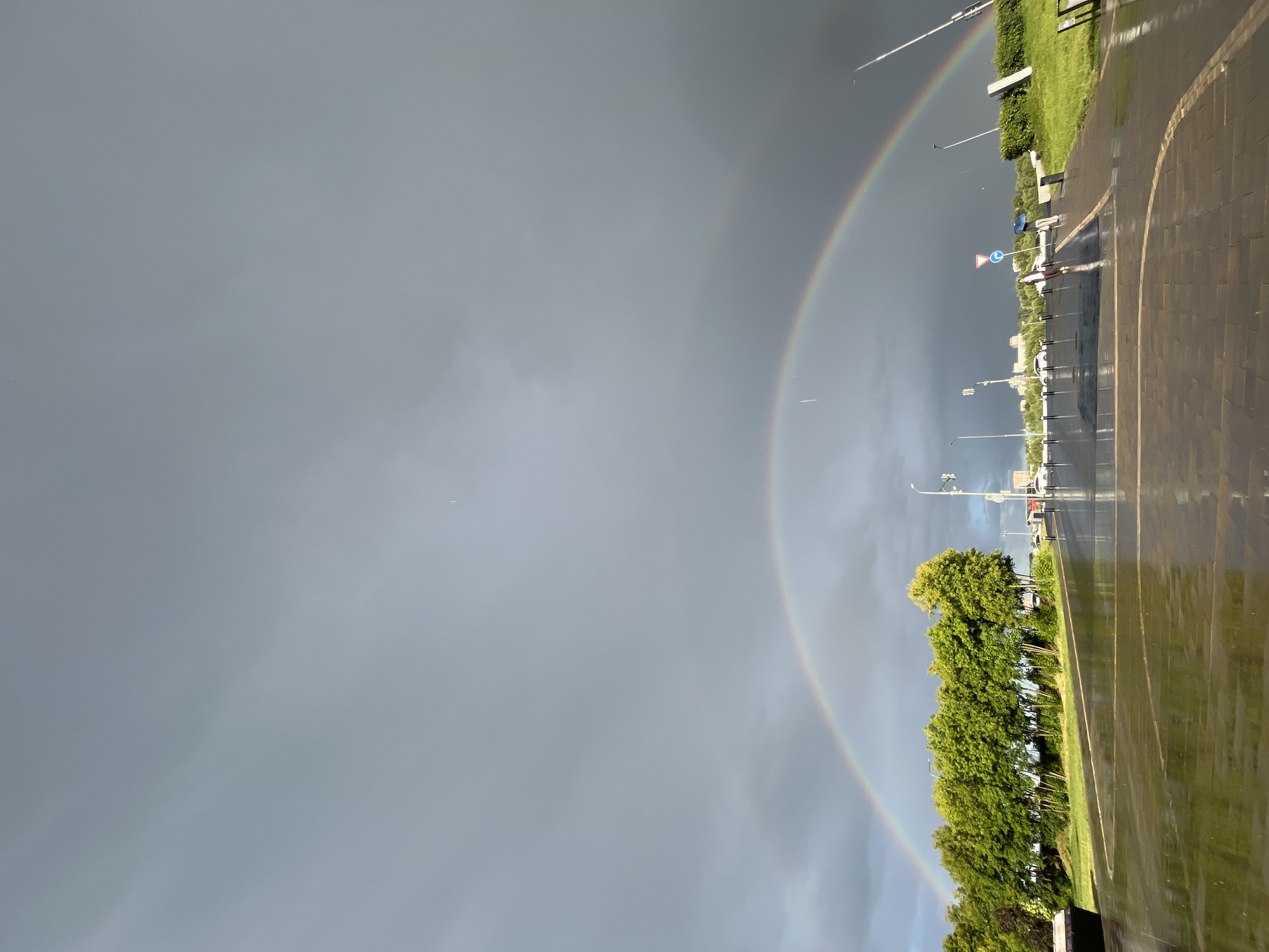 The media file depicts a scene with a prominent double rainbow arching across a gray, overcast sky. The image appears to be taken from a slightly elevated perspective, looking down and across a wet, paved area that resembles a parking lot. The paving stones are dark and reflect the subdued light.
To the right of the paved area, a vibrant green grassy verge runs alongside a line of lush green trees. Beyond the trees, a cityscape with buildings and streetlights is visible in the distance. Several cars are parked in the foreground and midground.
In the midground, near the trees, a few people are visible. One person is holding a blue umbrella, suggesting that it might have recently rained or is still drizzling. Another person, possibly a child, is walking with an adult. There are traffic signs visible, including a circular red and white sign.
The overall mood is one of a post-rain atmospheric scene, enhanced by the striking double rainbow. The wet ground and the presence of the umbrella indicate recent or ongoing precipitation. The time of day is likely late afternoon or early evening, given the quality of the light. The location cues and the specific appearance of the buildings and street furniture suggest Nizhny Novgorod, Russia. There is no discernible text in the image.