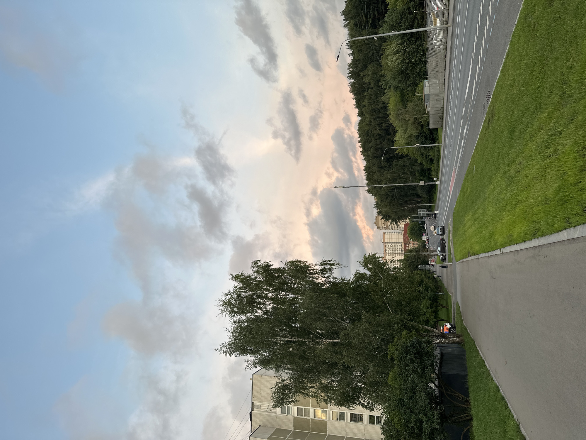 An eye-level, full shot captures a sidewalk running alongside a busy road in Moscow, Russia. The sky above is a gradient of light blue to pale pink, dotted with soft, scattered clouds. The road is a four-lane asphalt expanse, marked with white dashed lines and a red lane divider. On the right side of the road, a dense line of green trees forms a natural barrier. Beyond the trees, a light-colored fence and a few parked cars are visible. Streetlights with modern designs stand tall along the roadside.
On the left side of the image, a multi-story building with beige siding and numerous windows stands prominently. In front of the building, a large, leafy birch tree dominates the scene, its branches reaching towards the sky. A paved sidewalk runs parallel to the road, separated by a strip of vibrant green grass. Several pedestrians are scattered along the sidewalk, walking in various directions. A white and orange vehicle, possibly a service vehicle, is parked near the curb. The overall atmosphere suggests a calm evening in a suburban part of the city.