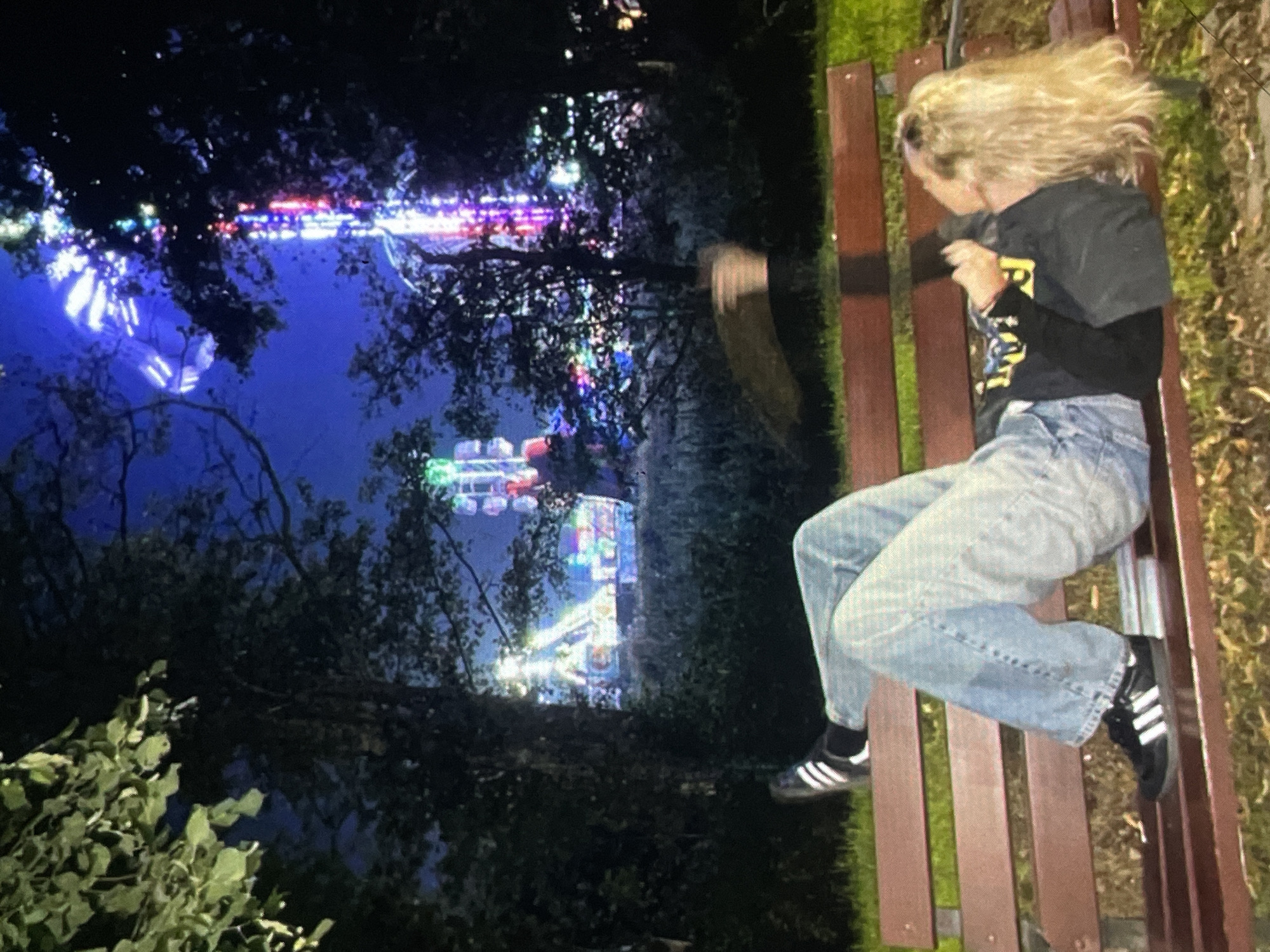 In Harmanli, Bulgaria, at night, a person is lying on a wooden park bench, reclined and facing away from the camera. They are wearing light-wash denim jeans and black athletic shoes with white stripes. The person has blonde hair and is wearing a black t-shirt with yellow lettering. In the background, a brightly lit amusement park is visible, with various rides illuminated by colorful lights against a dark blue night sky. Trees partially obscure the view of the park. The ground around the bench is covered in grass and fallen leaves.