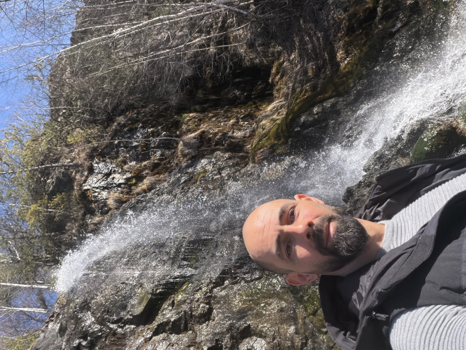 A bald, adult male with a dark beard is depicted in a close-up, chest-up selfie format, looking directly at the camera. He is wearing a grey ribbed long-sleeve shirt under a black puffer vest. Behind him, a waterfall cascades down a steep, rocky cliff face. The rock face is dark grey and brown, with patches of green moss and dry brown vegetation. Above and to the sides of the waterfall, deciduous trees with light-colored bark and bare branches, along with some evergreen conifers, are visible against a clear blue sky. The scene is outdoors, presumably near a natural water feature in Anton, Bulgaria.