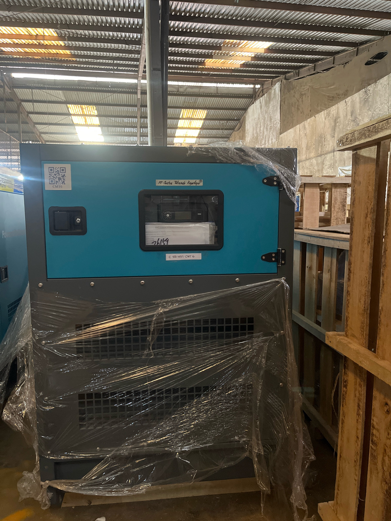 The image shows a generator unit, covered in plastic wrap, positioned inside a warehouse. The generator is primarily gray and teal, with a control panel visible on the front. Two QR codes are affixed to the teal section of the generator. The warehouse has a corrugated metal roof with natural light filtering through gaps. Wooden pallets are stacked to the right of the generator. The overall environment suggests a storage or distribution facility.