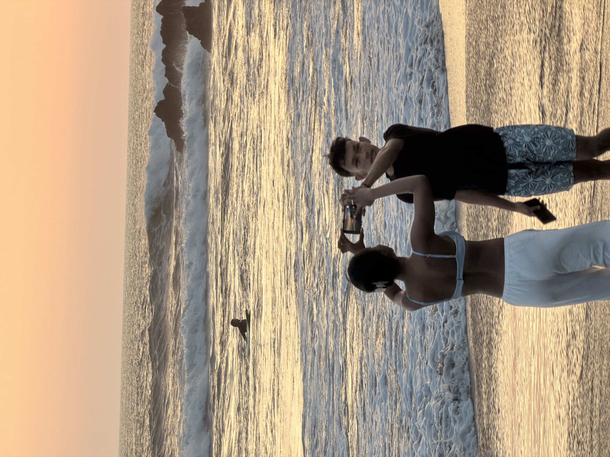 At sunset on the beach in North Kuta, Indonesia, a young woman wearing a blue bikini top and white pants stands with her back to the camera, holding a smartphone to take a photo. Next to her, a young man in a black t-shirt and patterned shorts holds a smartphone and appears to be taking a photo as well. In the distance, a surfer is visible on a surfboard in the ocean. Waves are crashing on the shore, and rocks are visible in the water. The sky is a soft orange, indicating the sun is setting over the ocean.