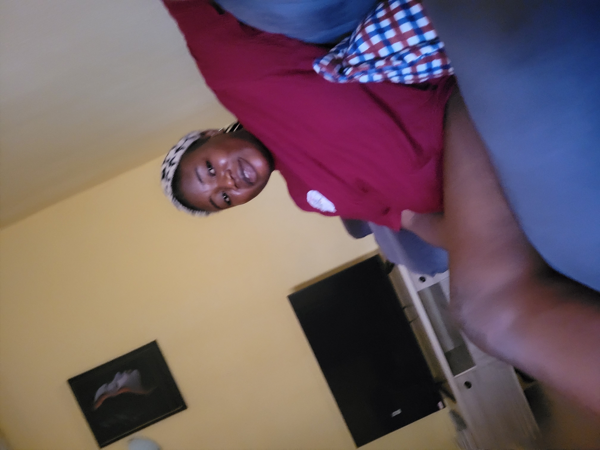 A full-body, slightly blurred, medium shot captures a young Black woman sitting on a sofa in a dimly lit room. She is wearing a maroon t-shirt, a patterned headband, and appears to be looking at the camera with a slight smile. Her left arm is extended upwards and outwards. A checkered bag sits on the sofa next to her. In the background, a black flat-screen television is mounted on a white entertainment stand against a yellow wall. A framed picture of a woman's profile is visible on the wall above the television. The scene suggests a casual indoor setting, likely a living room, located in Suleja, Nigeria.