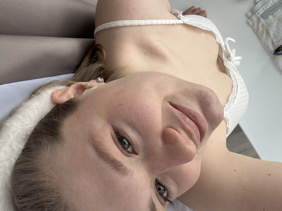 A young adult female is depicted in a high-angle, upward-looking portrait. She has fair skin, green eyes, and light brown hair, which is partially concealed by a white fuzzy headband. A small, clear, round stud earring is visible in her right earlobe. She is wearing a white ribbed camisole with lace trim and a small bow at the neckline. Her expression is a subtle smile, with her gaze directed towards the camera. The background consists of draped grey fabric, possibly curtains or upholstery. In the lower right foreground, a flat white surface is visible, bearing a partially seen folded white and yellow striped cloth, possibly a towel, and a dark, indistinct object. This scene is located in the city of Kazan, Russia.