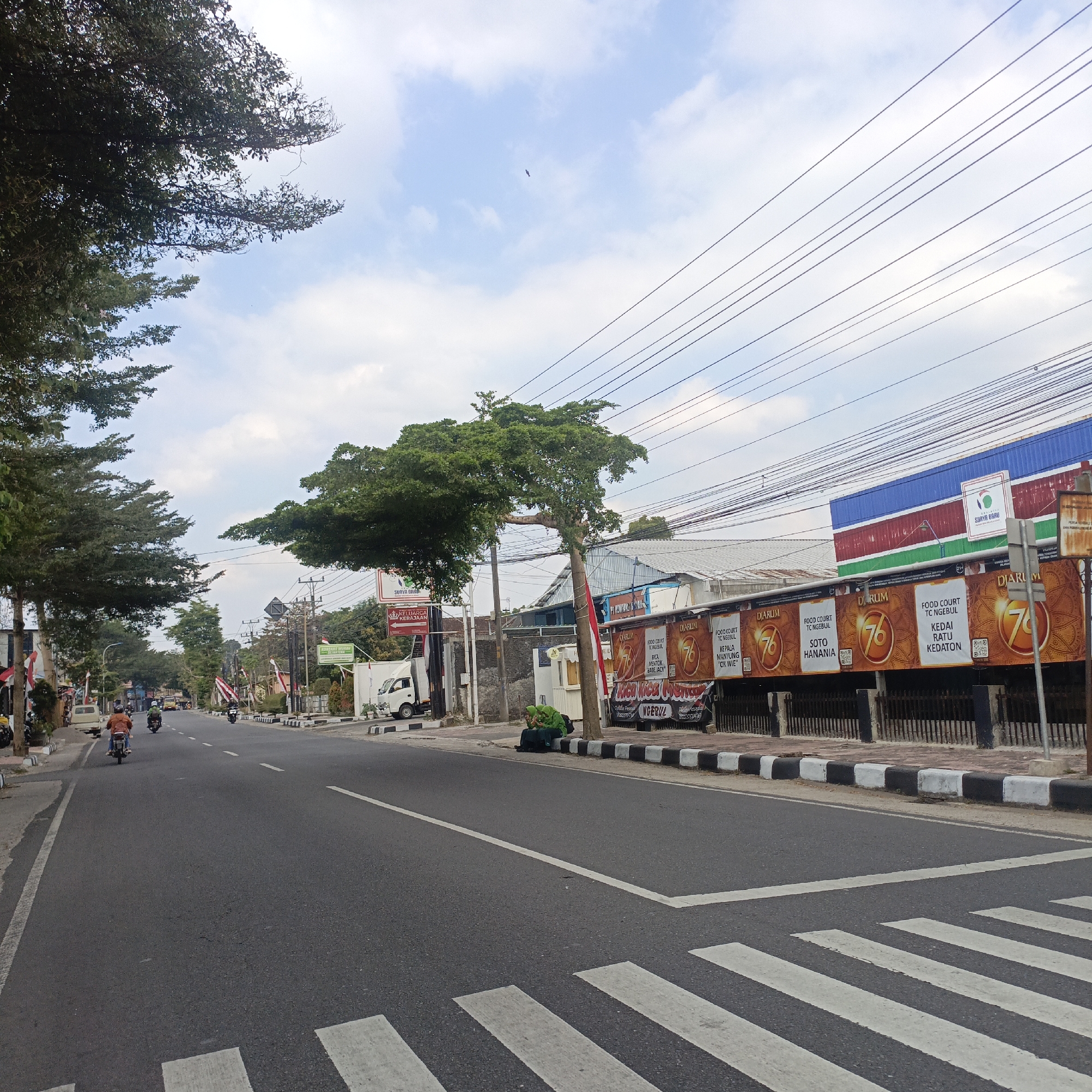 The image shows a street scene in Salatiga, Indonesia. A paved road runs through the center of the frame, with motorcycles and a white truck visible. The road is lined with trees on the left and buildings on the right. The buildings display advertisements, including ones for "DIARUM" and "FOOD COURT". A pedestrian crossing is marked on the road. The sky is partly cloudy. Two individuals are seated on the sidewalk in front of a building. Overhead, power lines are strung across the sky.
