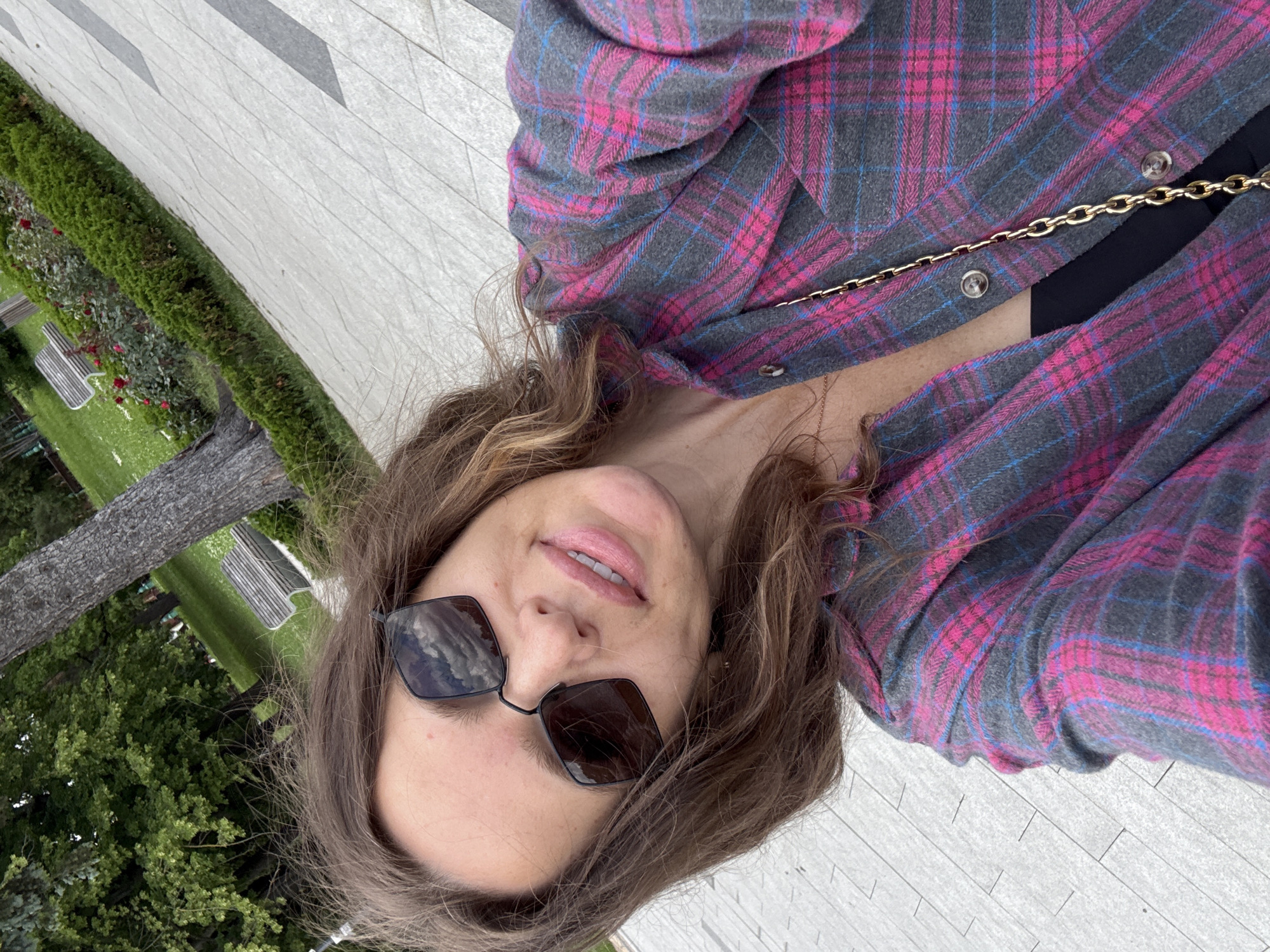 A woman takes a selfie outdoors, possibly in a park in Moscow, Russia. She is wearing a pink and gray plaid shirt over a black top, with a gold chain strap visible across her chest. She has on dark, square-shaped sunglasses that reflect the sky. Her brown hair is loose and slightly windswept. The background shows a paved walkway, green grass, trees, and a park bench. The overall lighting suggests an overcast or diffused daylight condition.